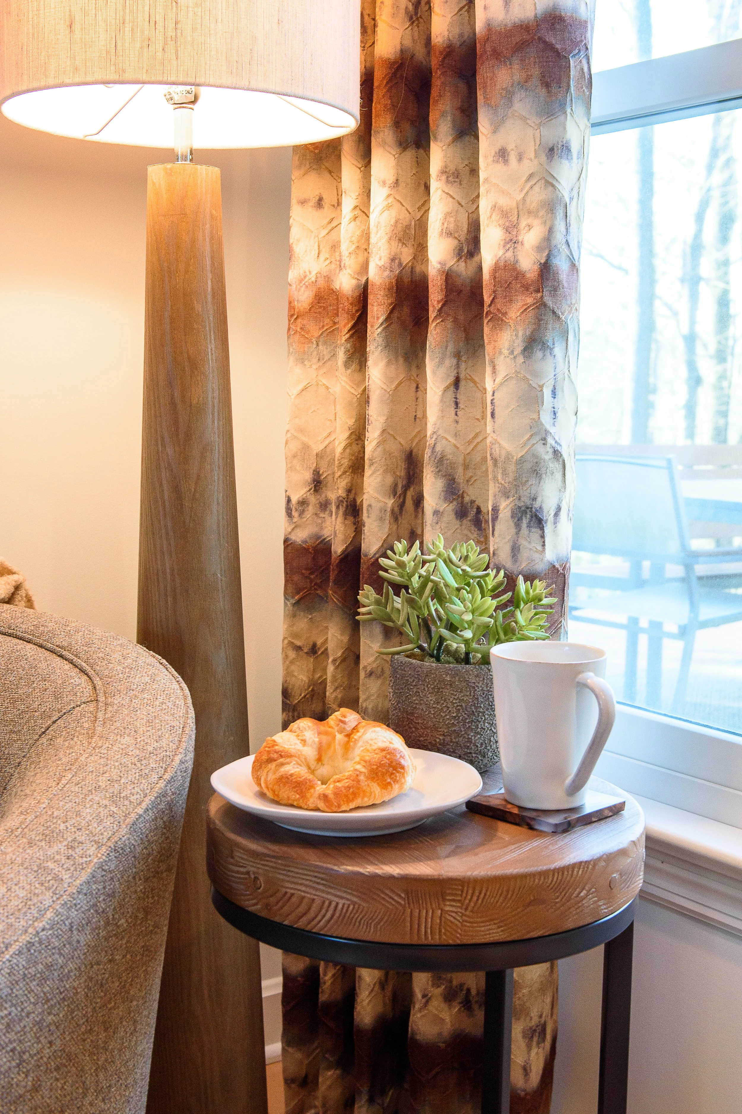 Modern end table with a croissant on a white plate, a white coffee mug, and a succulent in a gray stone pot, set in front of cream, brown, beige, and gray‑blue curtains with an outdoor view behind them.