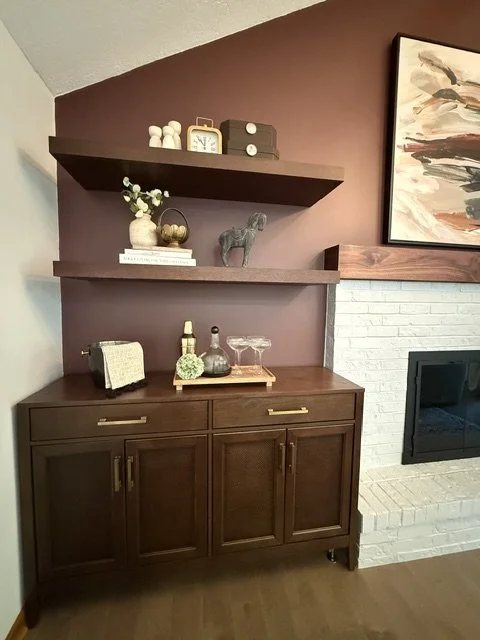 Interior of a living room with a dark wooden cabinet, decorative shelves with plants and decor, a colorful abstract painting, and a fireplace with a white brick surround.