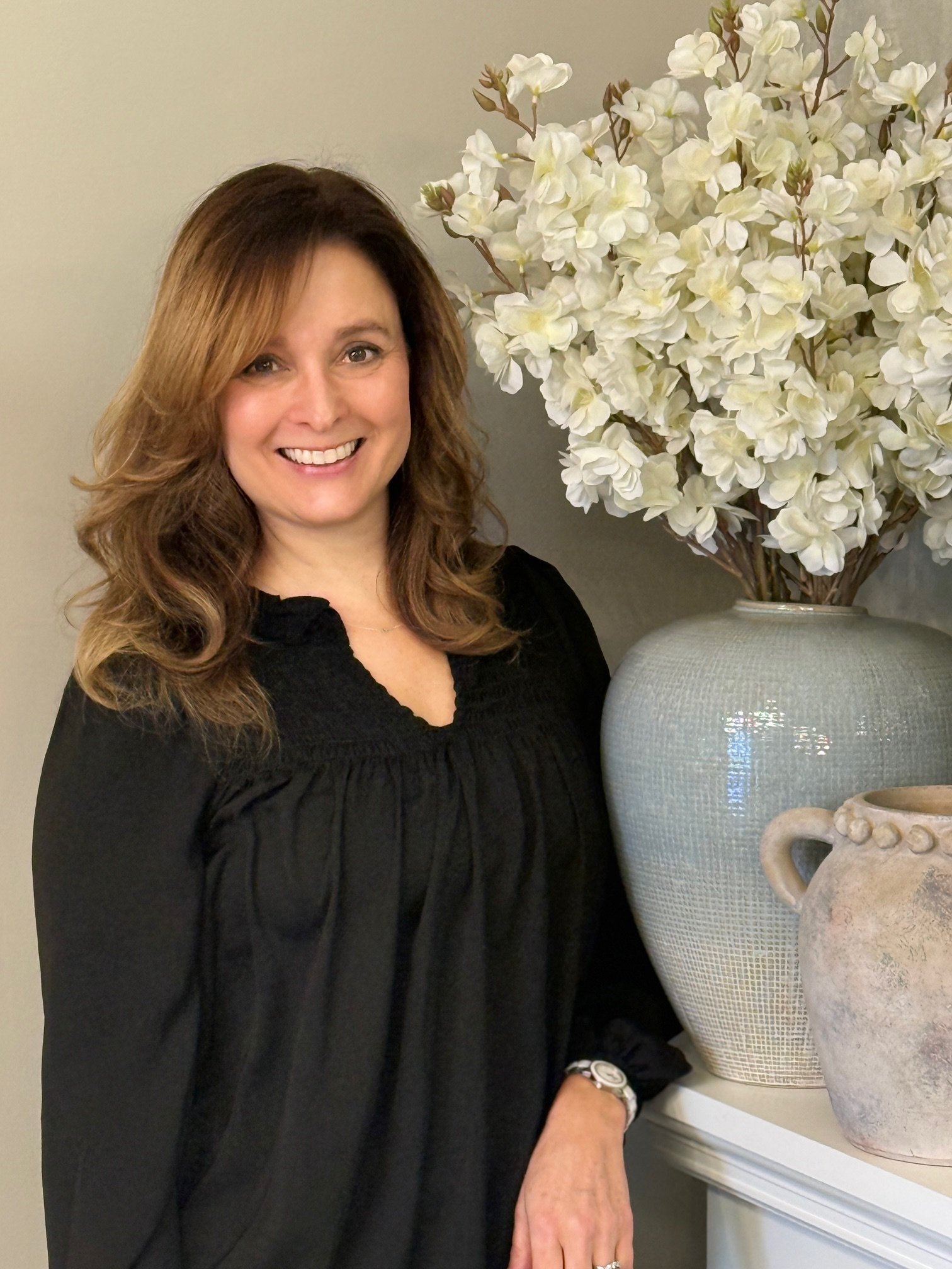 Portrait of Gina Wilkes wearing a black blouse, with white flowers and light-colored vases in the background.