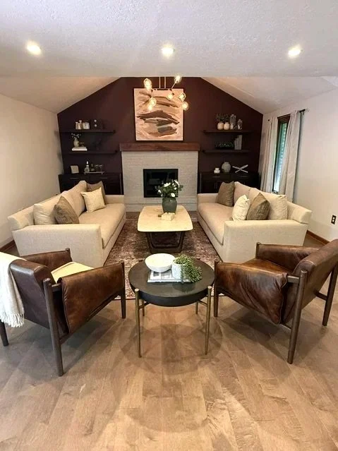Living room with two beige sofas facing each other, two brown leather chairs, a coffee table with a plant and books, a fireplace with a marbled mantel, and wall shelves with decor, in a room with hardwood floors and a vaulted ceiling.