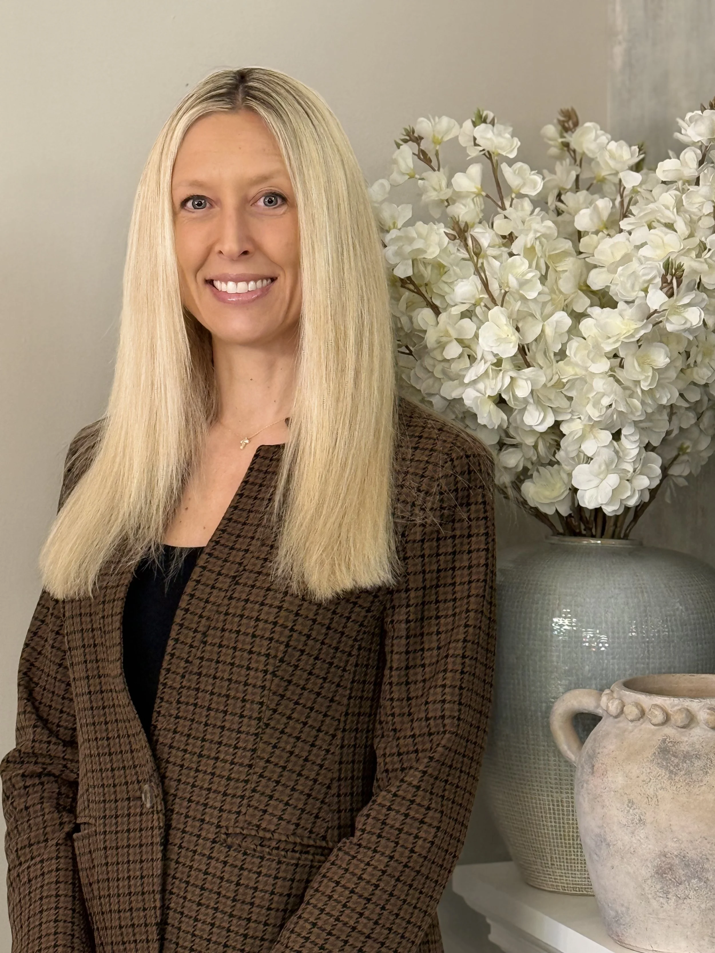 Portrait of Erin Kovach wearing a black shirt with a brown and black houndstooth print blazer, with white flowers and light-colored vases in the background. 