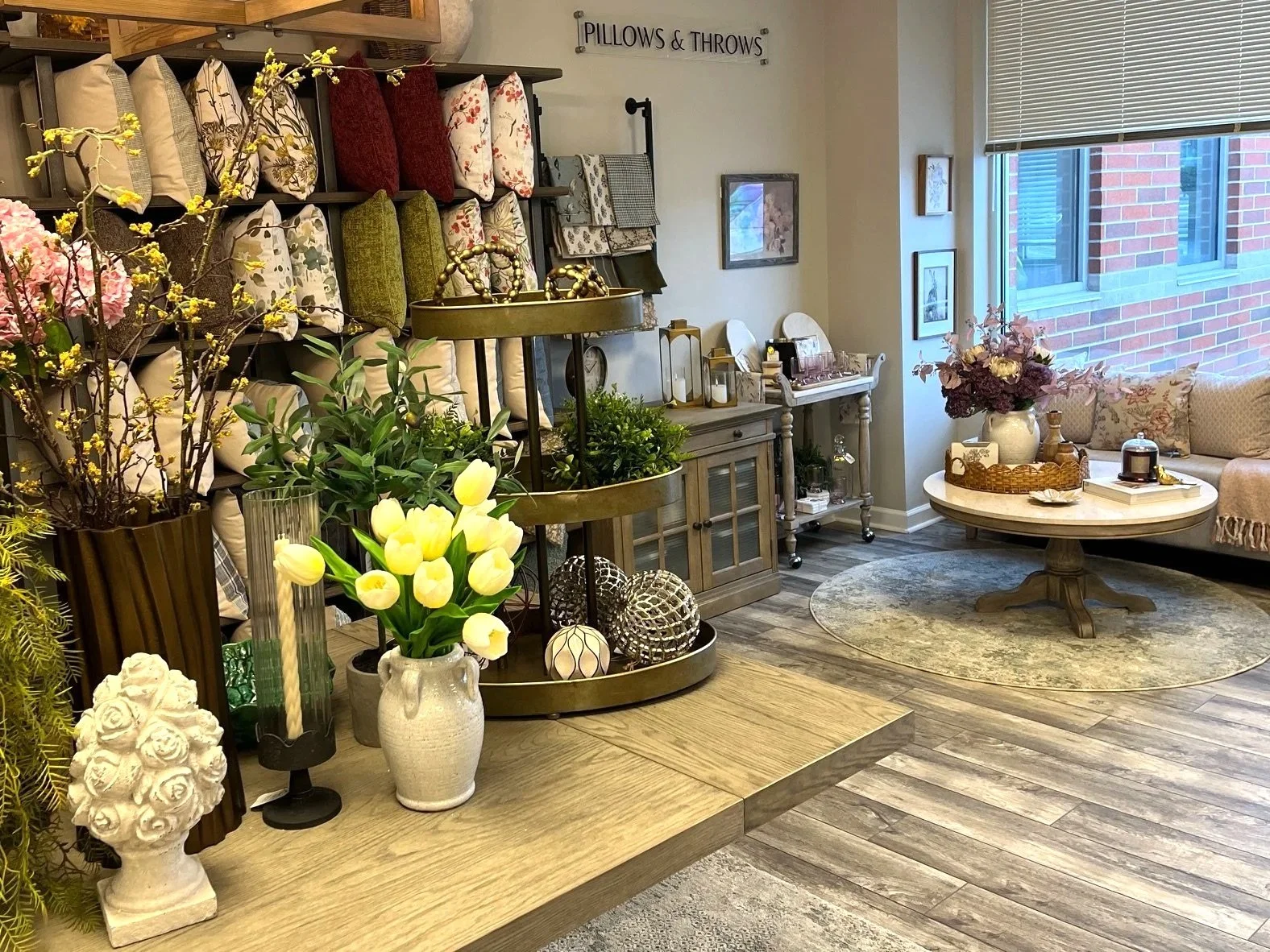 A living room with a display of decorative pillows, flowers, and home décor items, including a round coffee table with floral arrangements, and a window with blinds.