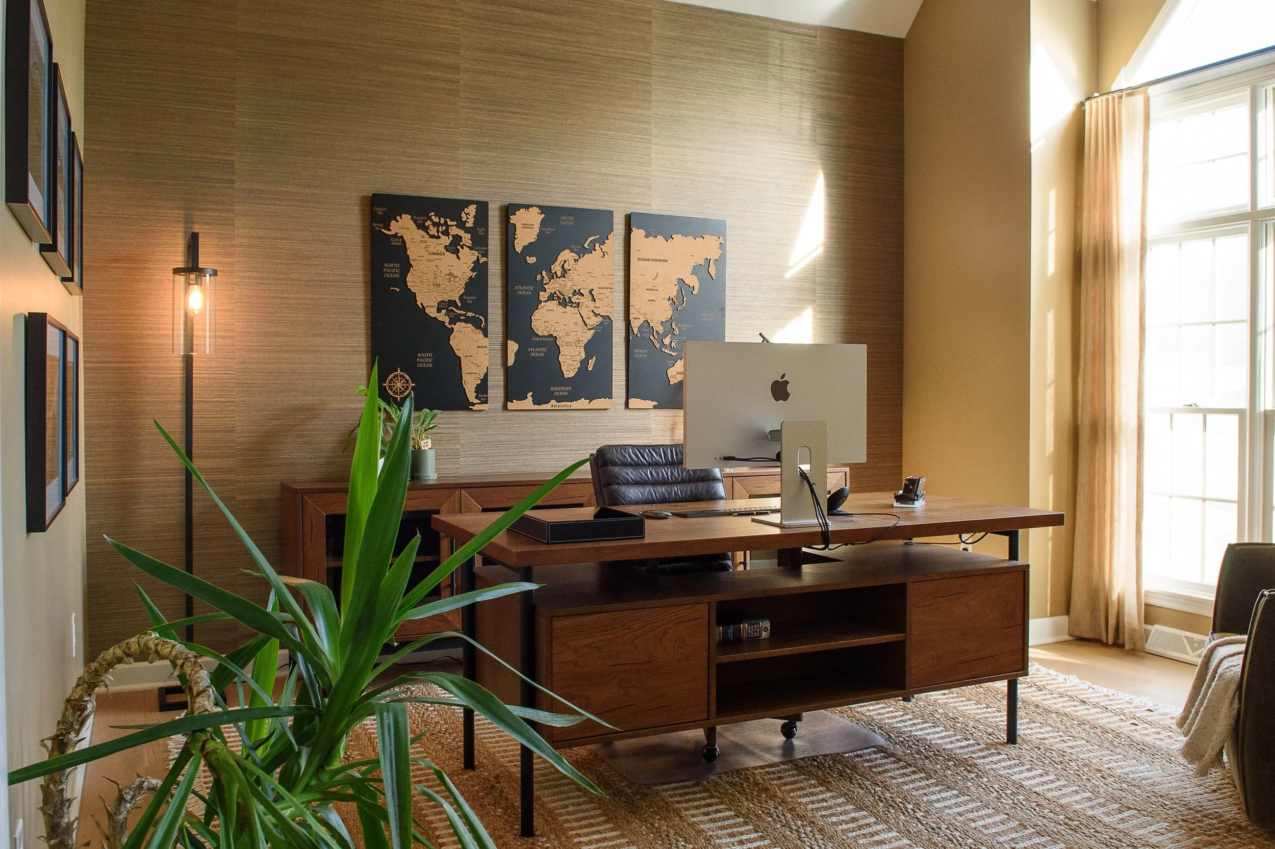 An office with a wooden desk and a black leather chair, a large iMac computer on the desk, a map of the world on the wall, window curtains, and a potted plant in the foreground.