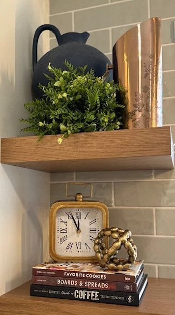 Decorative black teapot, green potted plant, and a large gold-colored book on a wooden shelf against a tiled wall, with a clock, a gold sculpture, and stacked books below.