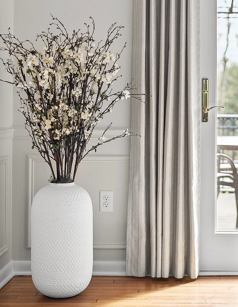 A large white vase with textured surface holding tall branches with white flowers, placed next to a window with patterned curtains and a wooden floor.