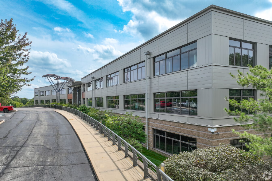 Modern commercial building with large windows, adjacent parking area, sidewalk, landscaped greenery, and a blue sky with clouds.
