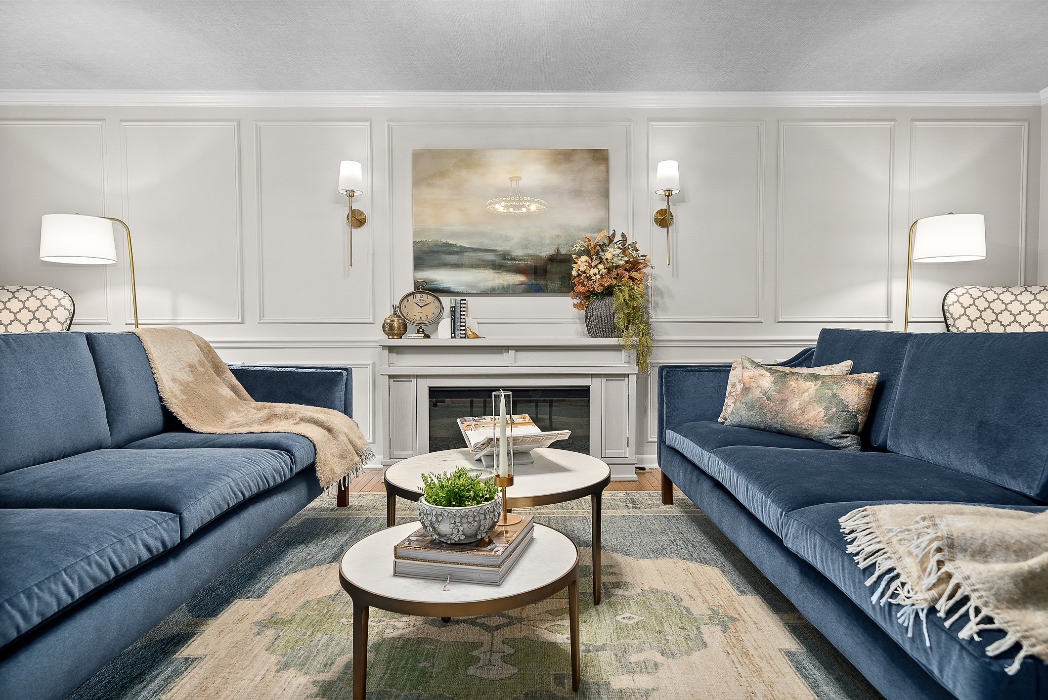 Living room with two blue sofas, a fireplace with decorative items on the mantle, and wall-mounted lamps. A painting hangs above the fireplace, and there are two coffee tables with decor and books, on a patterned area rug.