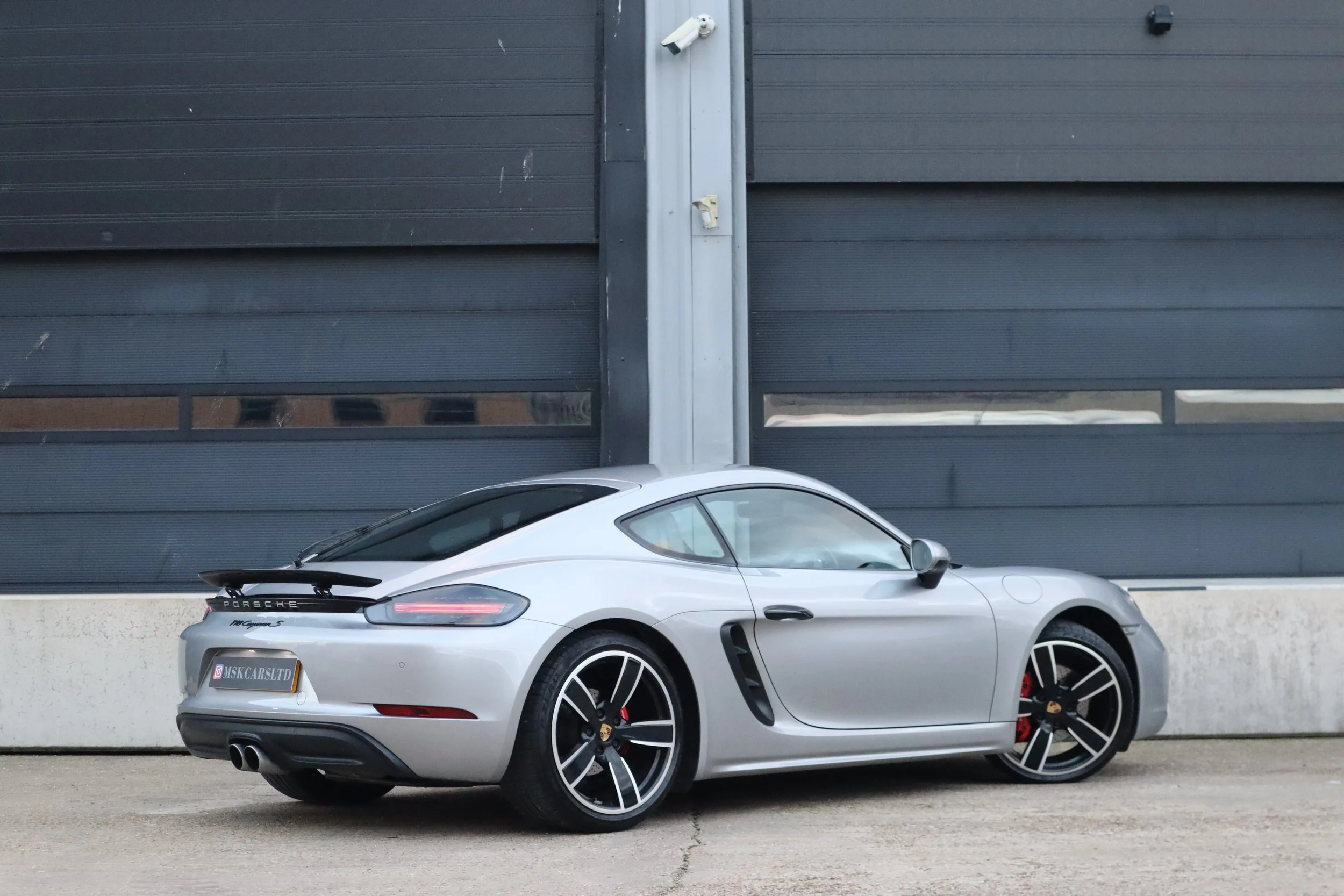 Silver Porsche 911 coupe parked in front of a black garage door with two windows, on a concrete surface.