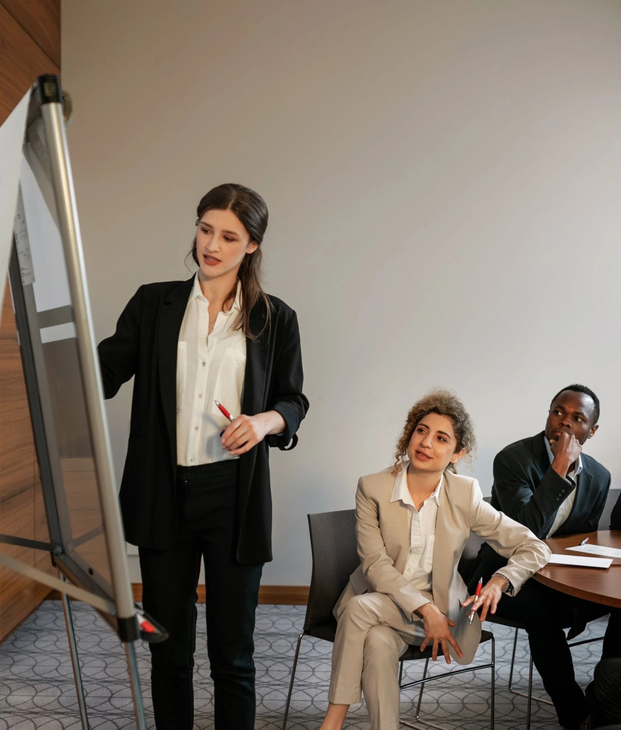 A woman in a black blazer and white shirt is standing and holding a red pen while speaking to a group. Three individuals are seated at a table, listening; they are a woman in a beige blazer, a woman with curly hair, and a man in a dark suit. A flip chart stand is visible on the left side of the image.