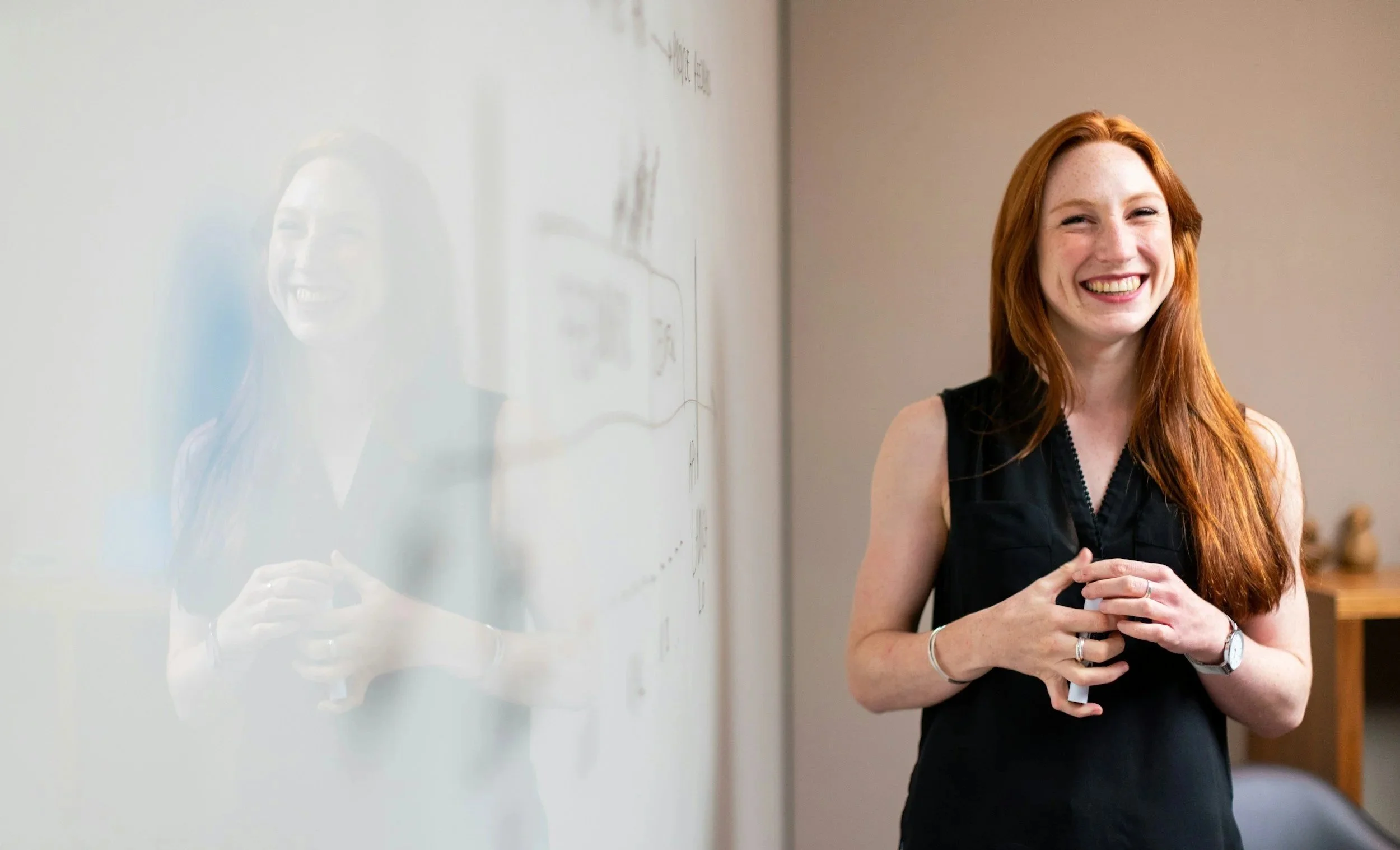 A woman with long red hair and a black sleeveless top standing in front of a whiteboard, smiling and holding her hands together.