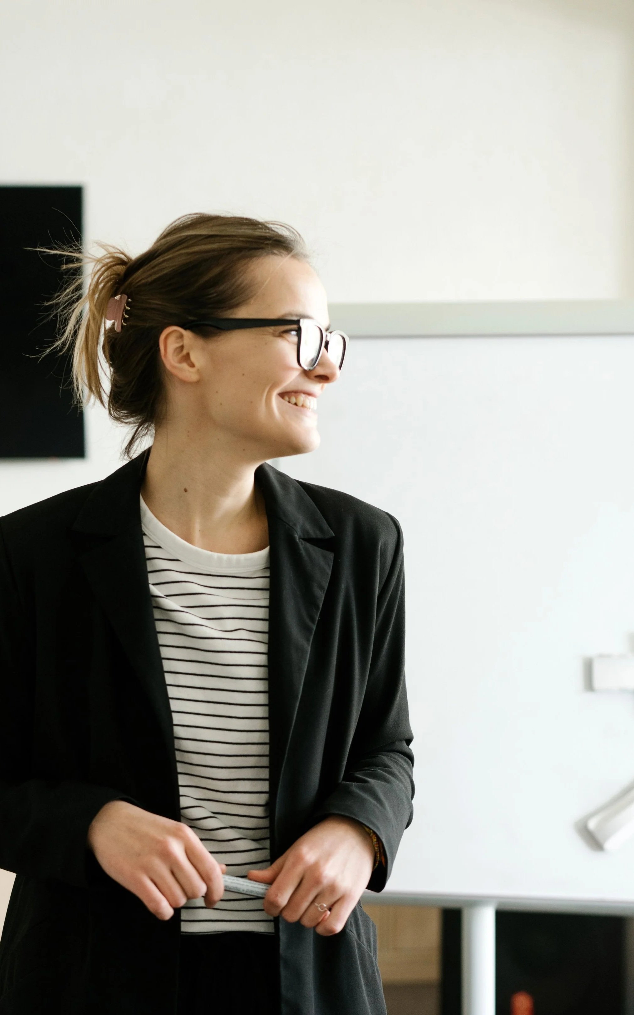 A young woman with glasses, tied back hair with a pink hair clip, smiling, wearing a black blazer over a striped shirt, standing in a bright, modern office or classroom setting.