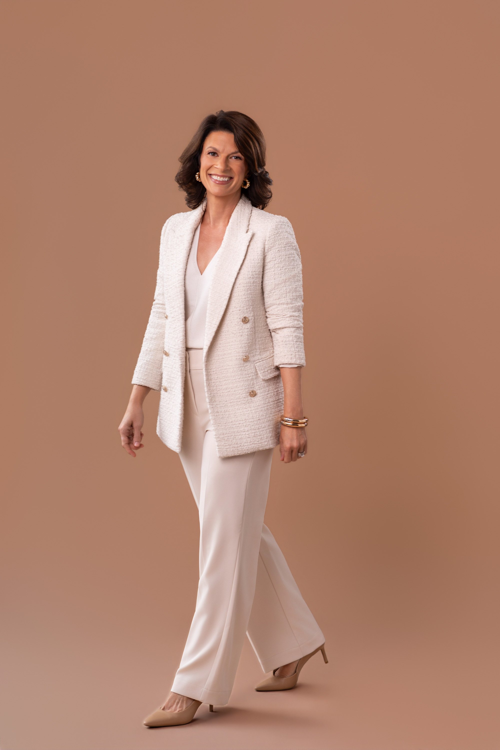 A woman with dark shoulder-length hair, wearing a cream-colored textured blazer, white blouse, cream wide-leg trousers, beige high heels, gold earrings, and bracelets, smiling against a solid brown background.
