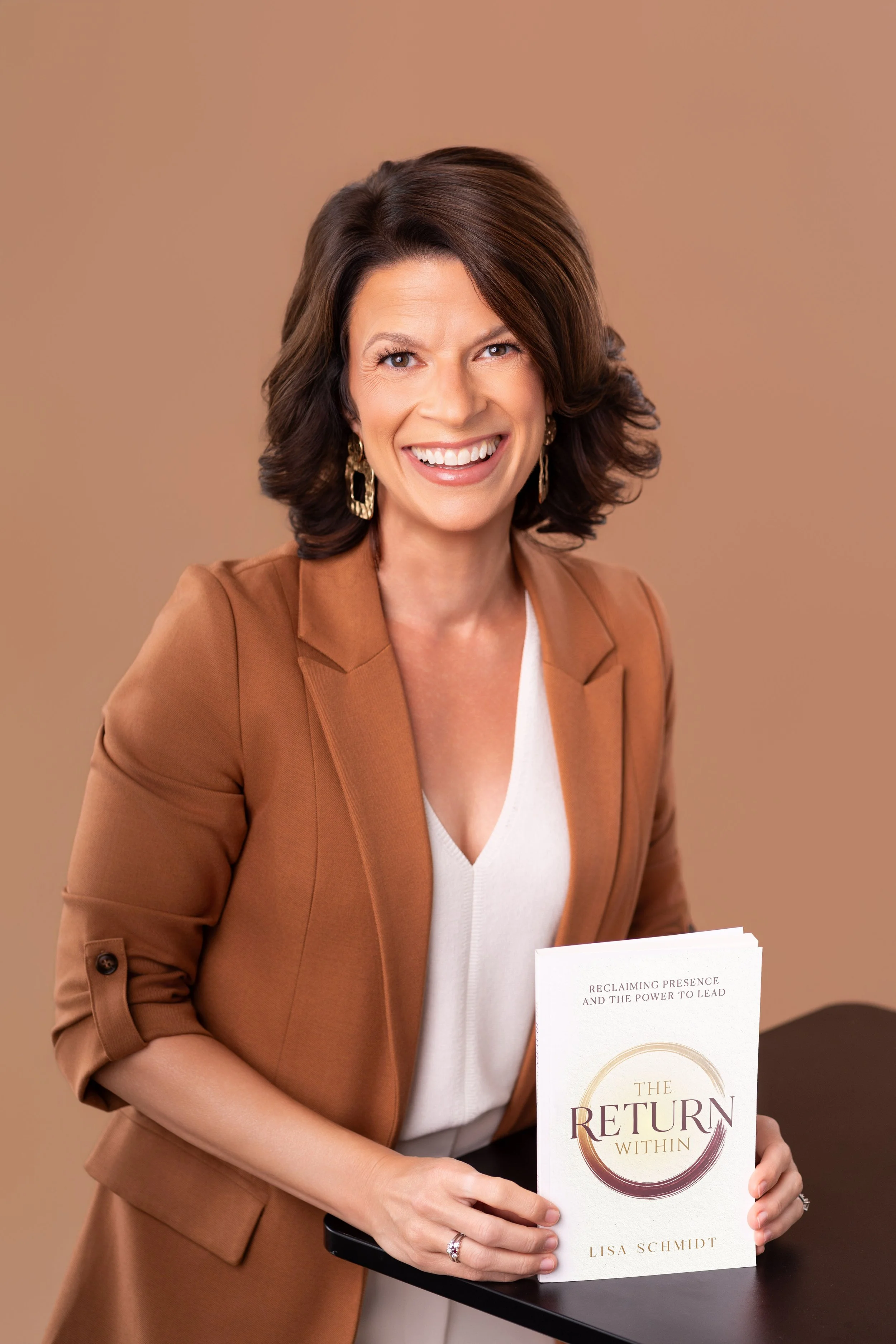 A woman with shoulder-length brown hair smiling, wearing a tan blazer over a white top, holding a book titled "The Return Within" by Lisa Schmidt, with a beige background.