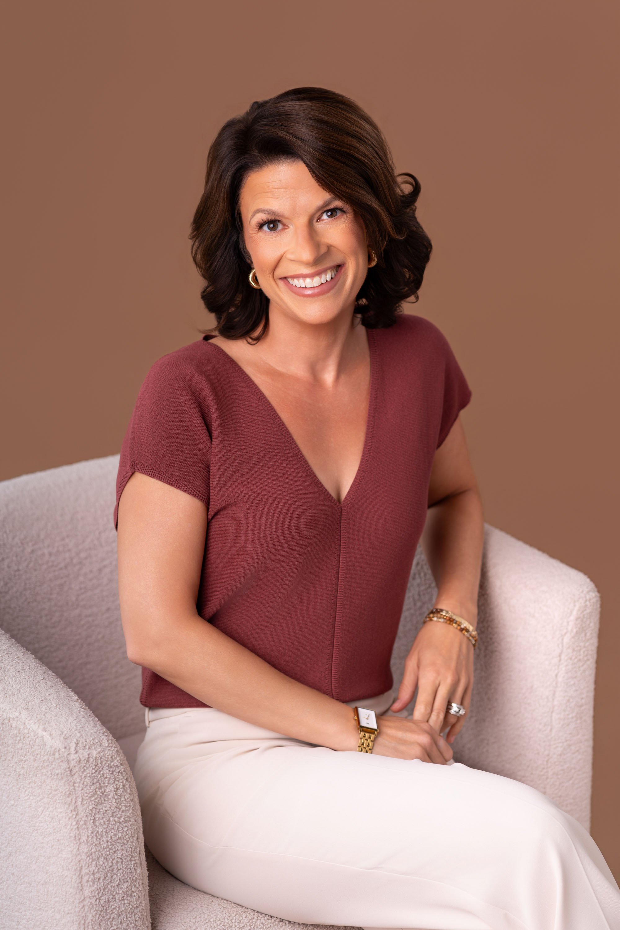 A smiling woman with dark brown hair styled in loose waves, wearing a maroon V-neck top, gold earrings, and jewelry, seated on a light beige upholstered chair against a solid brown background.