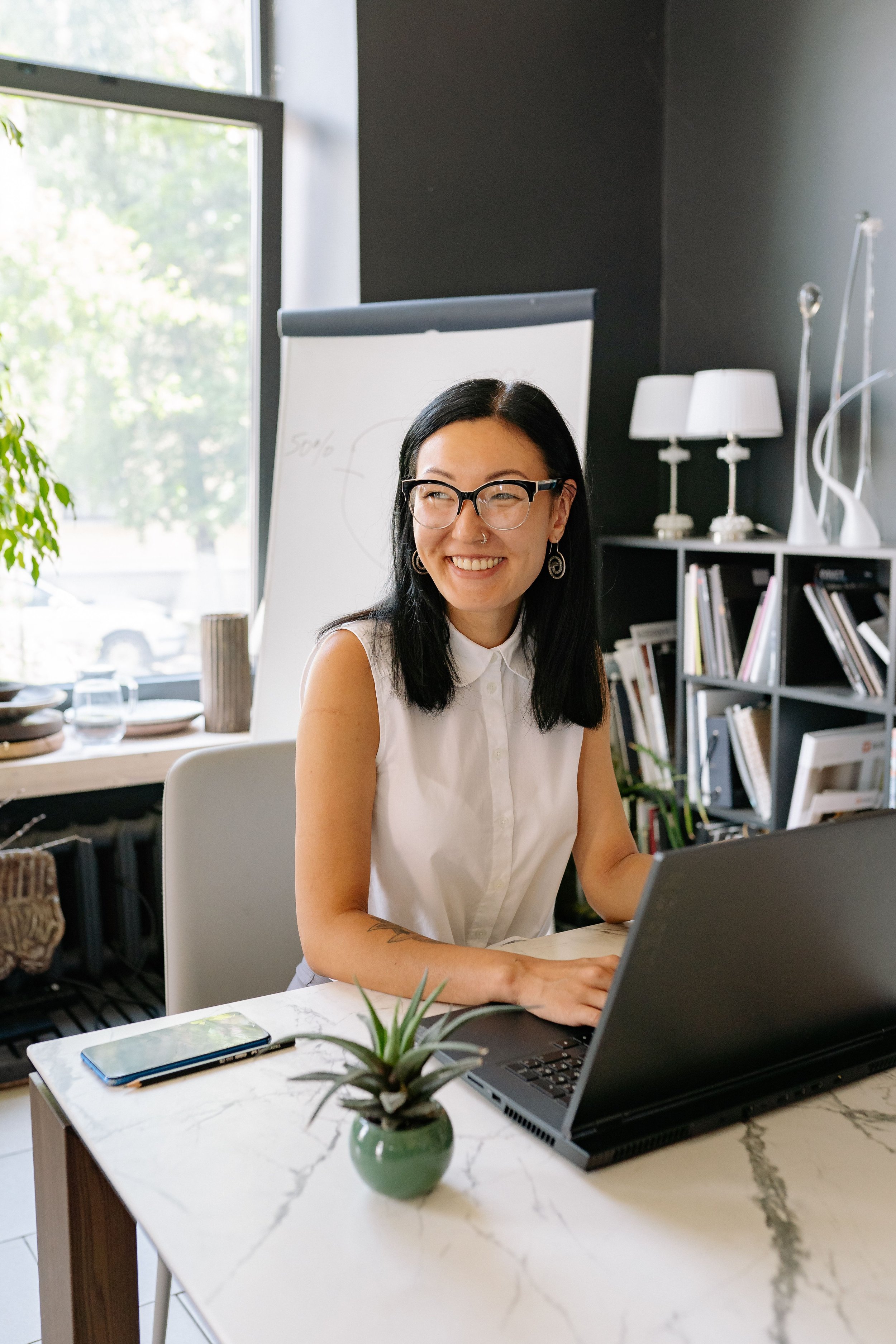 A woman with glasses and earrings smiling as she uses a laptop at a marble-top table in a modern, well-lit office or workspace.