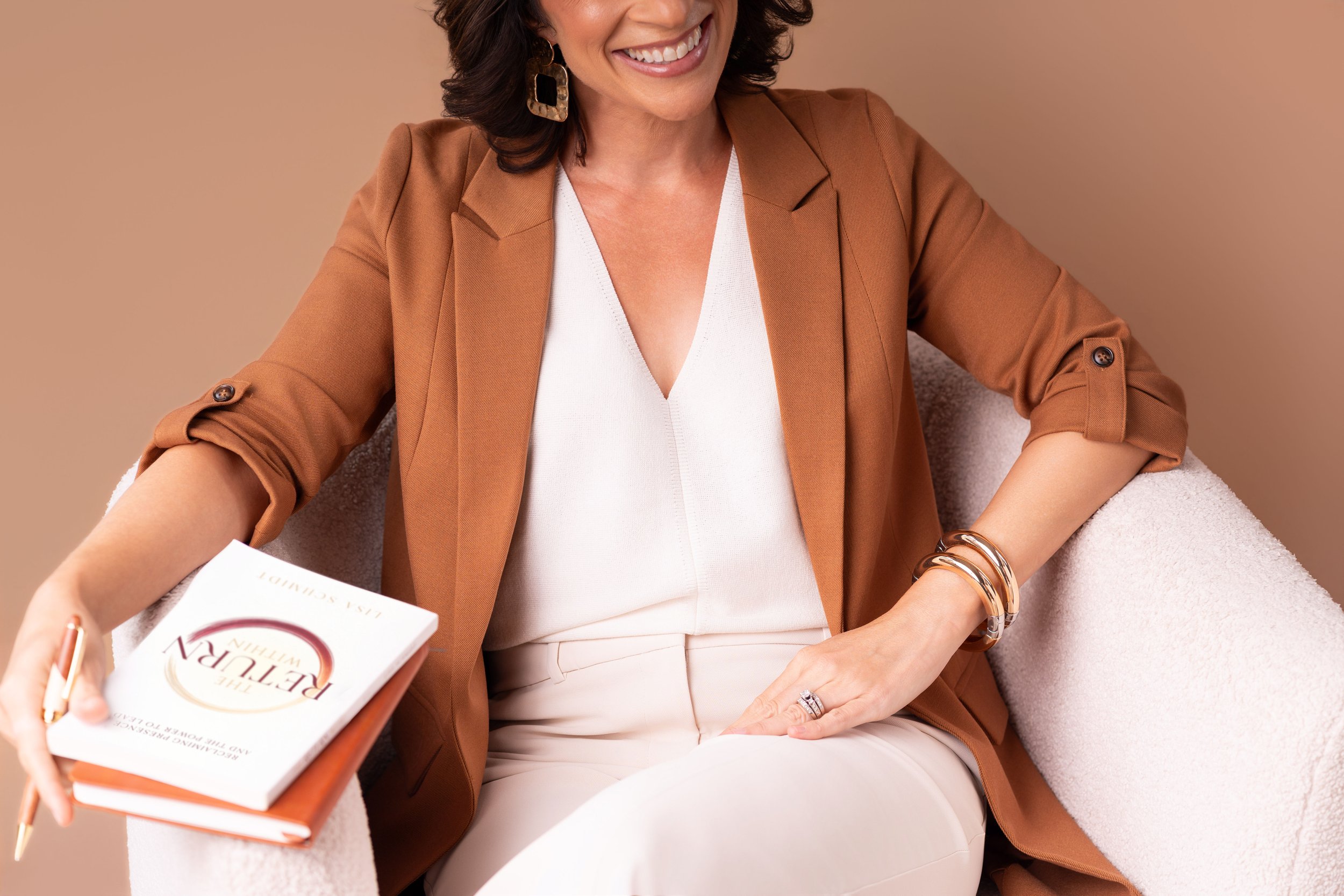 A woman with dark, wavy hair smiling and wearing a brown blazer, white top, and gold jewelry, sitting on a light-colored chair against a beige background, holding a book titled 'Return' and a pen.