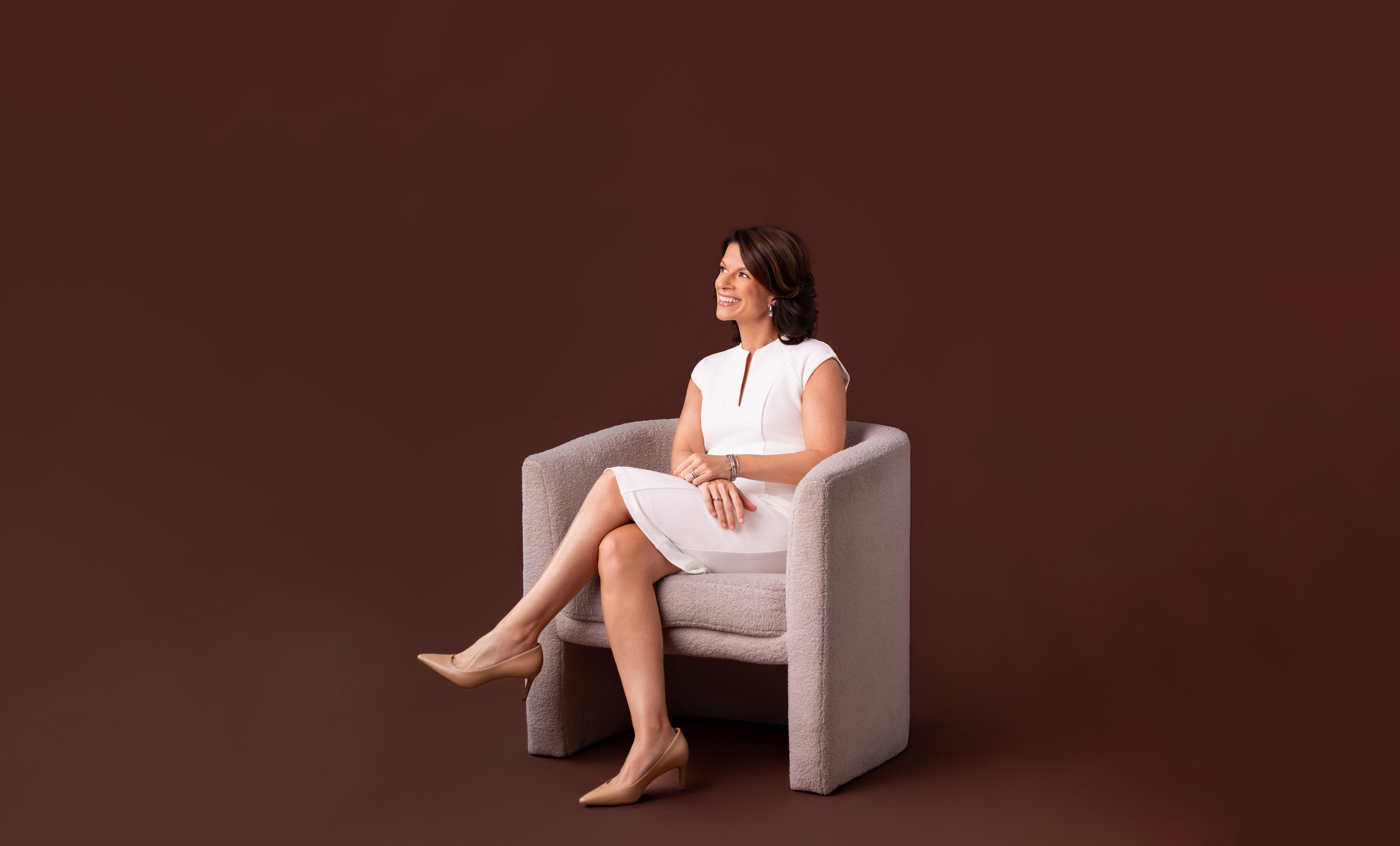 A woman with brown hair wearing a white dress sitting on a light gray armchair against a brown background, smiling and looking to the side.