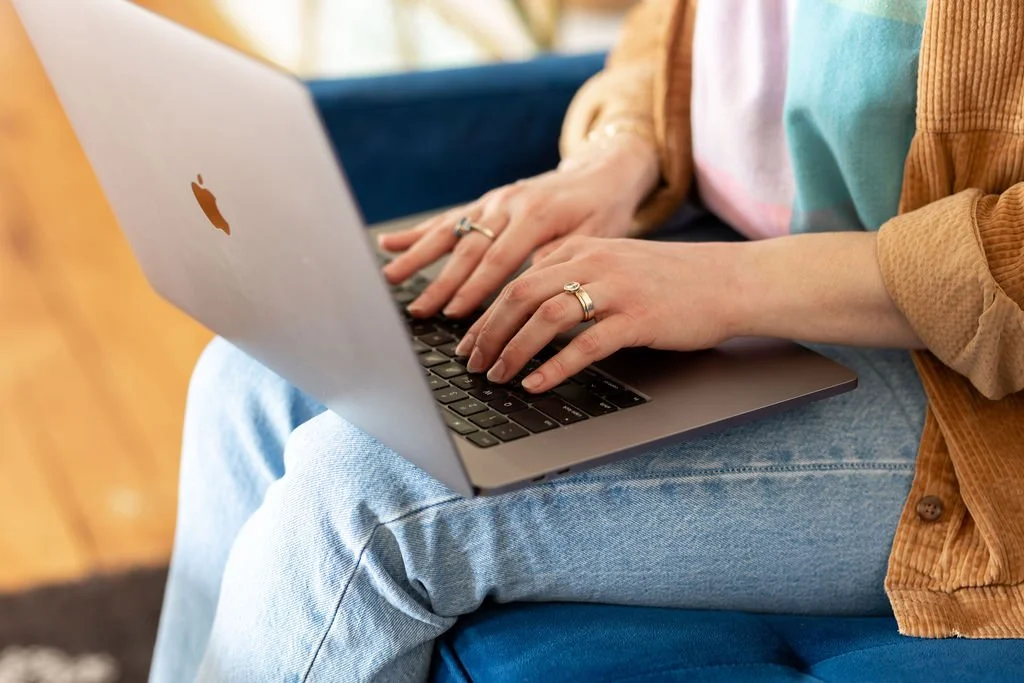 A close up shot of a white woman with jeans  a colored shirt and tan corduroy over shirt types on a silver Macbook laptop. She is wearing rings.