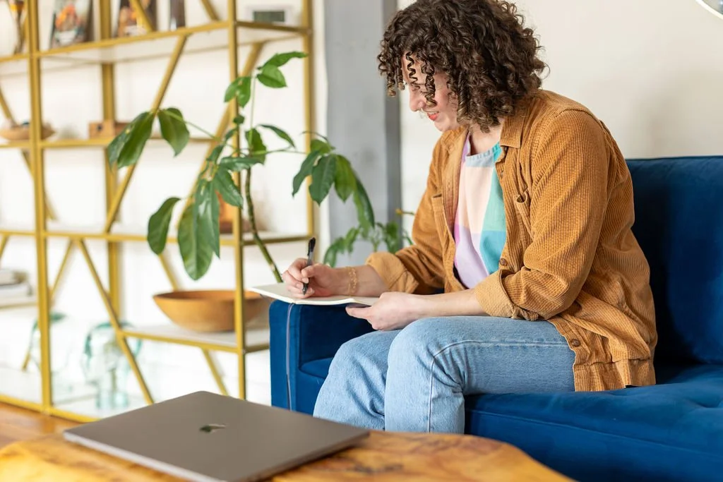 A white woman in her mid-thirties with chin length brown curly hair sits on a blue velvet sofa and writes in her journal. In front of her is a table with a silver Macbook laptop. She is wearing jeans, a colored undershirt, and a tan corduroy buttonup