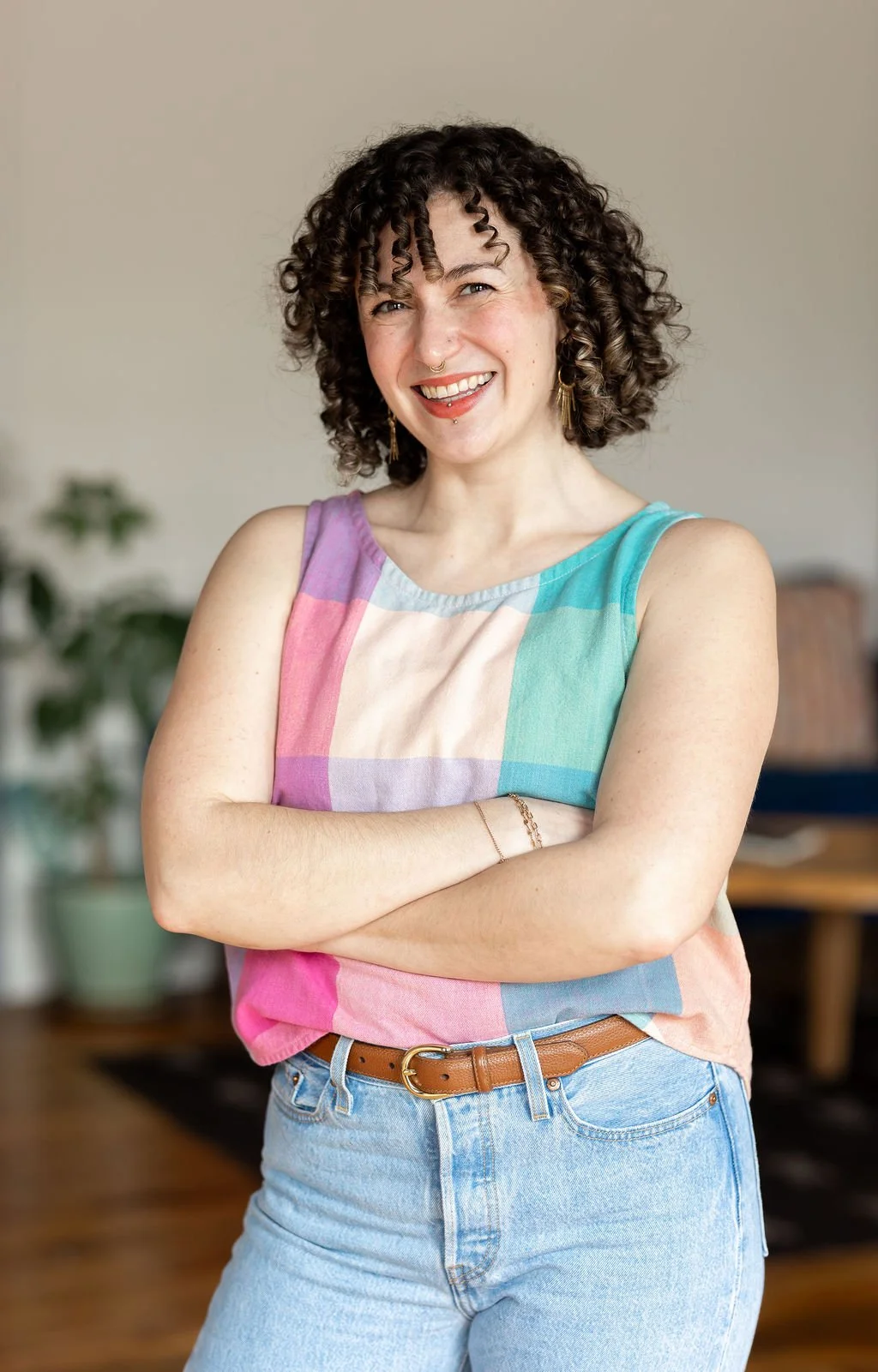 A white woman in her mid-thirties with facial piercings, chin-length brown curly hair, a pastel large-scale gingham check tank top, and her arms crossed.