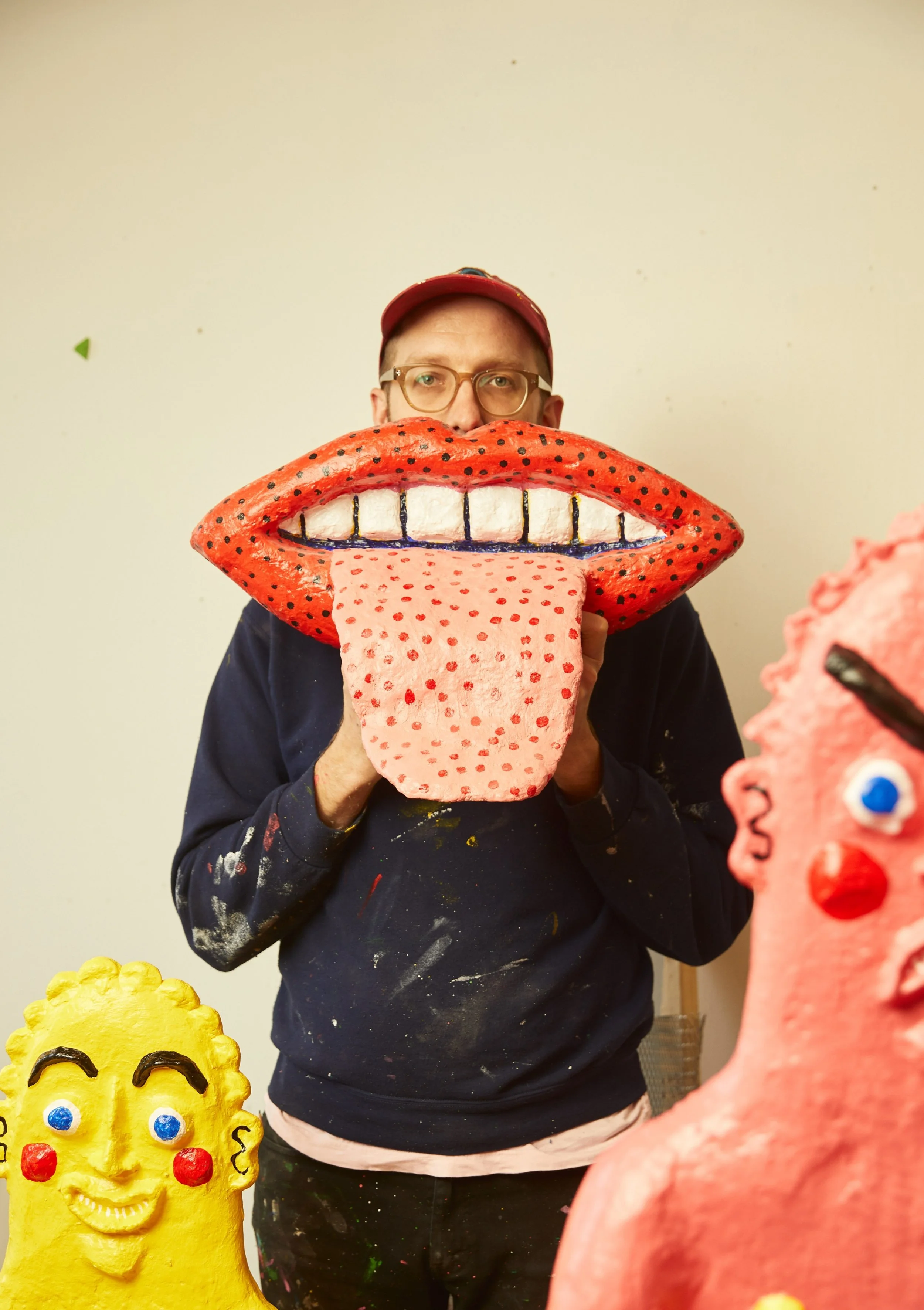 Person holding a large red lip-shaped mask with a tongue sticking out, surrounded by colorful, cartoon-like sculptures of faces.