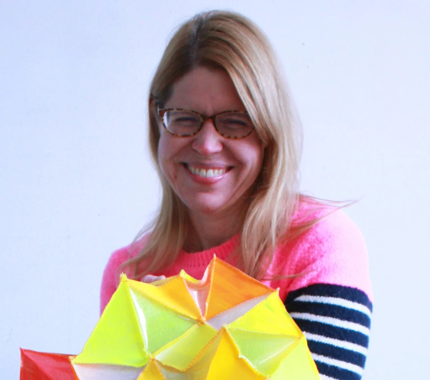 A woman with blonde hair and glasses smiling, holding colorful paper umbrellas.