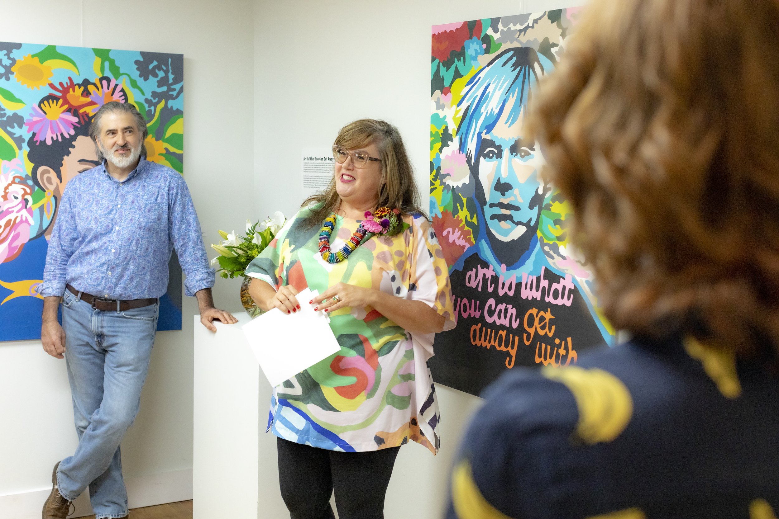 Three people standing in front of colorful portrait artwork at an art gallery opening. The woman in the center is wearing a vibrant, multicolored shirt with flowers and jewelry, holding a piece of paper, and speaking to the woman in the foreground. The man on the left leans against a white pedestal, smiling. The woman in the foreground has curly hair, wearing a dark shirt with yellow accents, and is listening to the woman.