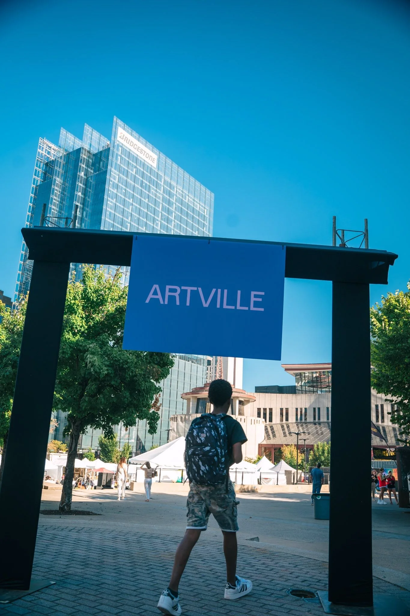 A young person with a backpack walking through an outdoor art festival with tents, trees, and modern buildings in the background.