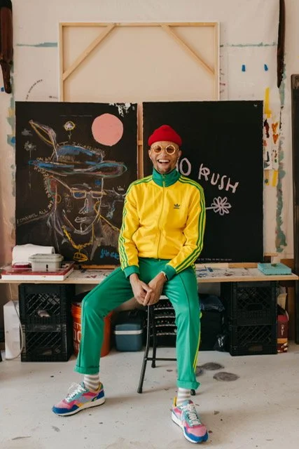 Person sitting on a chair in front of two blackboards with colorful drawings, wearing a yellow and green tracksuit, red beanie, pink sunglasses, and colorful sneakers, smiling.