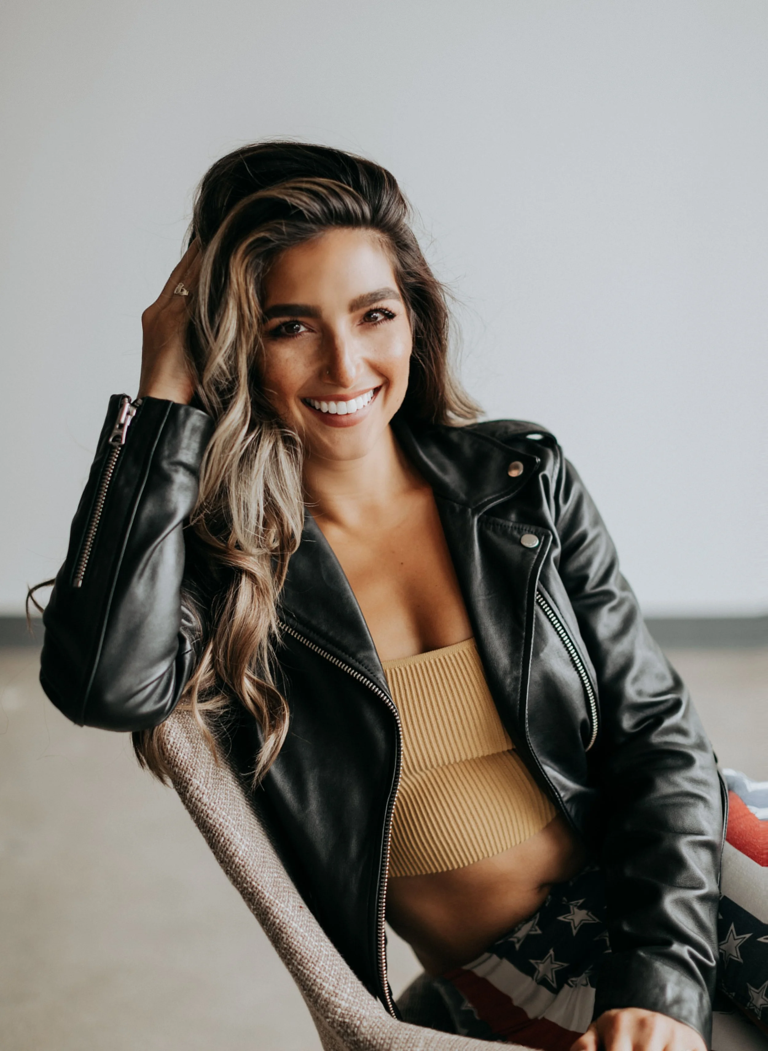 A woman smiling and sitting on a chair, wearing a black leather jacket, a beige ribbed crop top, and star-spangled shorts.