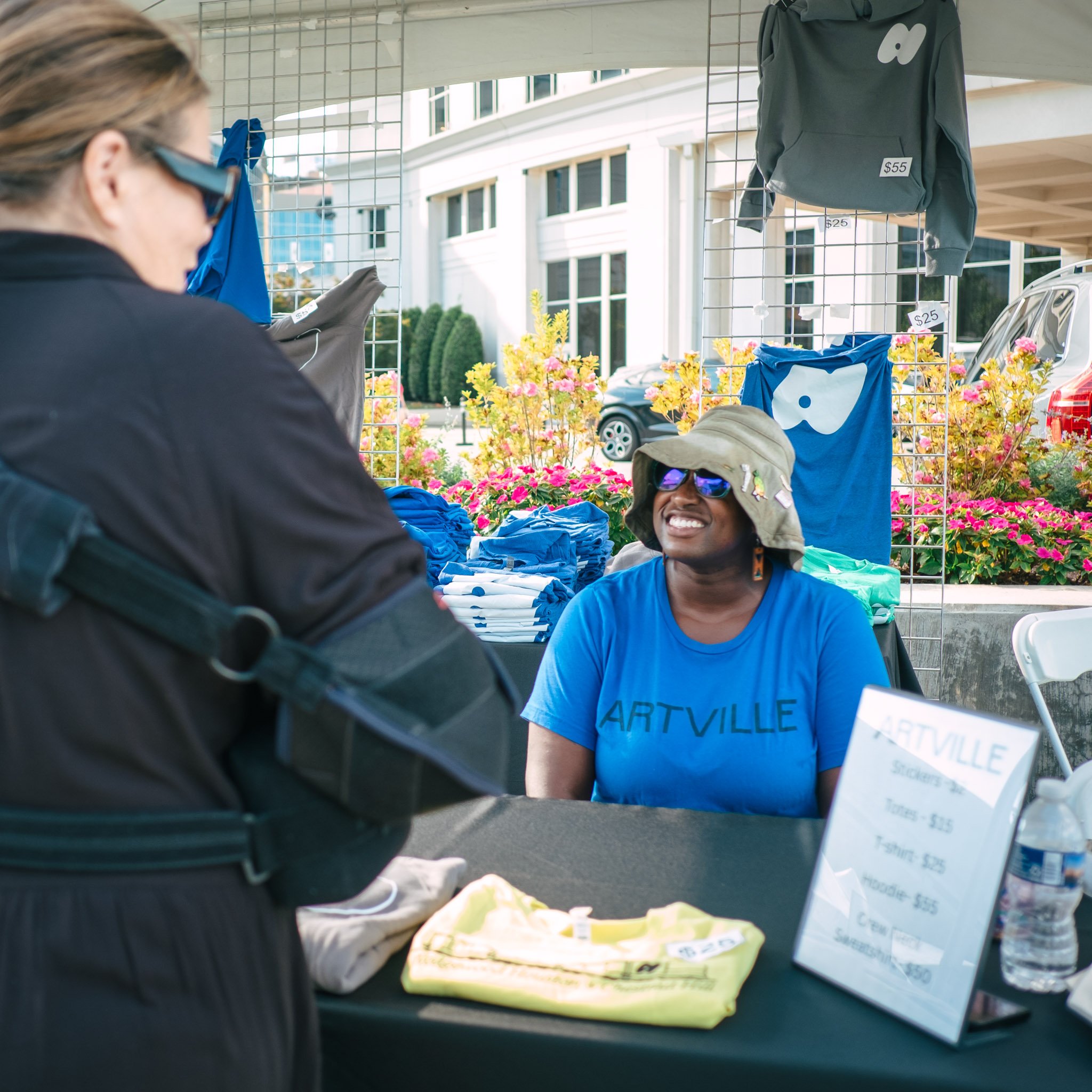 A woman with sunglasses and a blue t-shirt labeled "ARTVILLE" is smiling and looking at a table with baked goods at an outdoor market. There are signs with prices and stacks of baked items in the background.