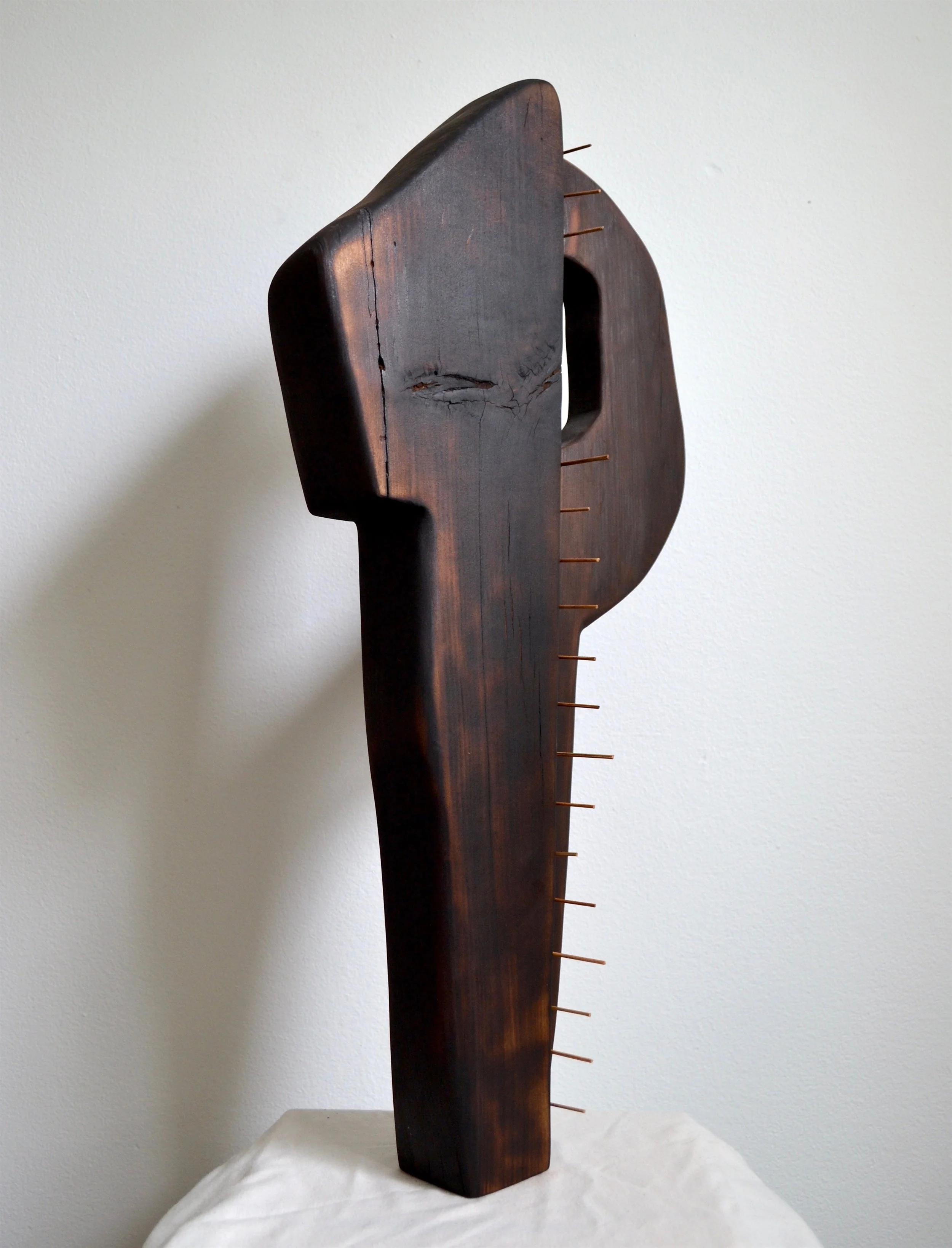A tall, dark wooden tribal mask sculpture with carved details and protruding nails or sticks, standing on a white cloth against a plain white wall.