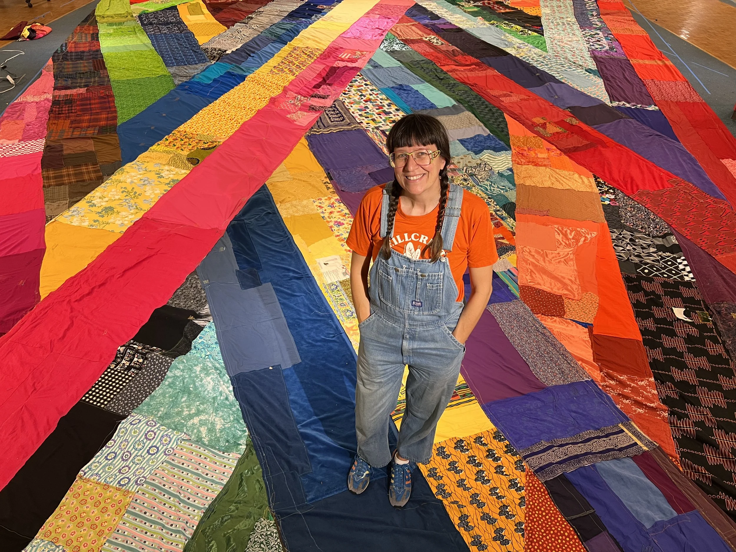 A woman with glasses, braided hair, wearing an orange shirt and denim overalls stands smiling on a large quilt made of various colorful fabric patches.
