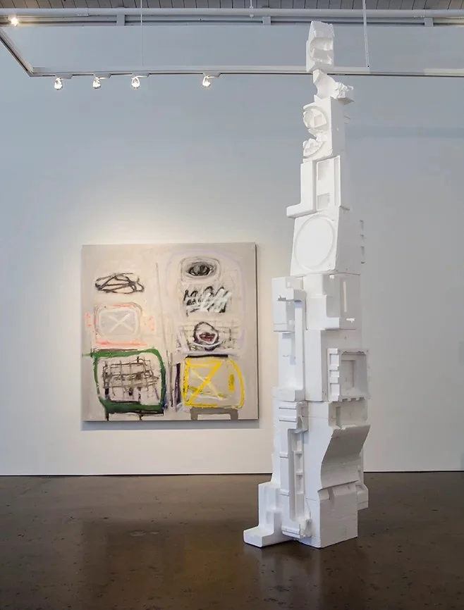 An art gallery featuring a tall white abstract sculpture made of stacked and carved block-like forms, with a modern abstract painting on the white wall in the background.