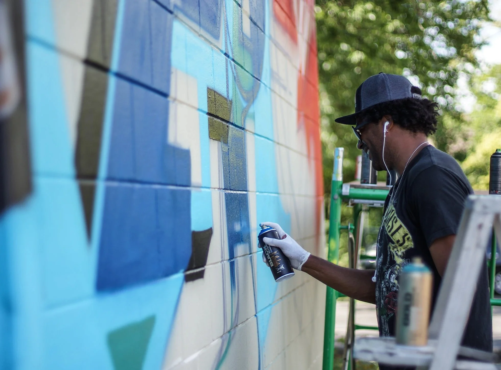 A man wearing a baseball cap, sunglasses, and gloves is spray painting a mural on an outdoor brick wall, standing on a ladder. Trees and sunlight are visible in the background.