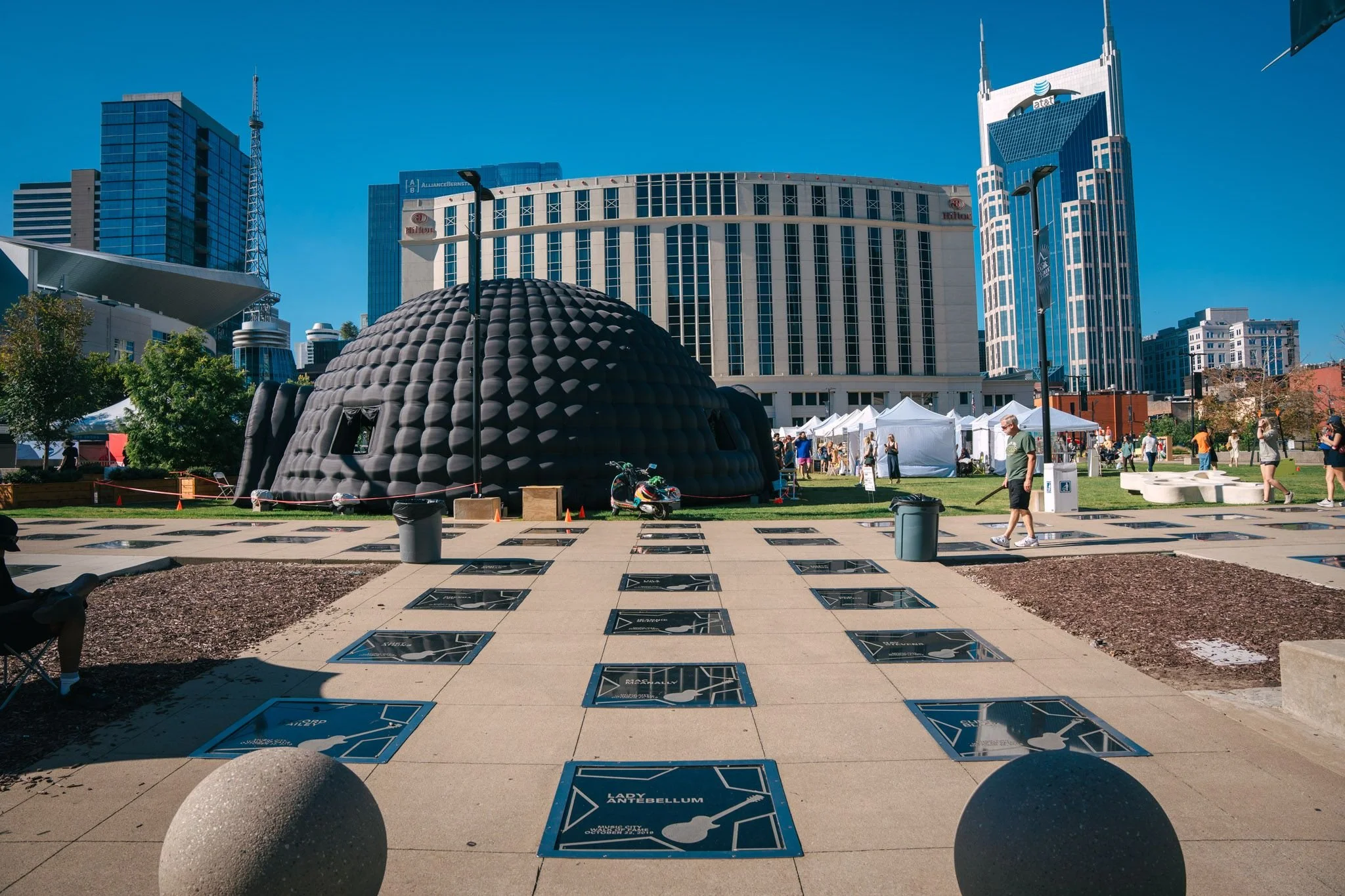 Cityscape with tall modern buildings, a domed black inflatable sculpture, and a small outdoor event with white tents and people walking on a sunny day.