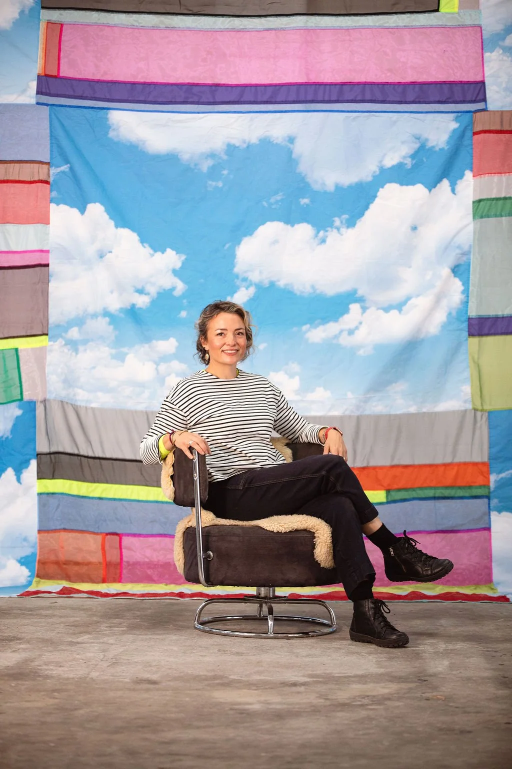A woman sitting on a chair in front of a studio backdrop that depicts a bright blue sky with white clouds. She is wearing a striped long sleeve shirt, black pants, and black boots, smiling at the camera.