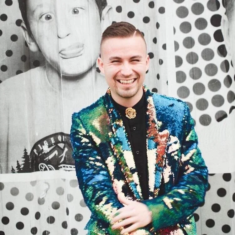 A smiling man in a colorful, shiny jacket standing in front of a black-and-white poster featuring a boy with his tongue out and a polka dot background.