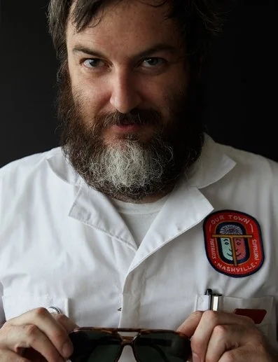 Man with dark hair and beard wearing a white shirt with a Nashville public safety badge, holding sunglasses against a dark background.