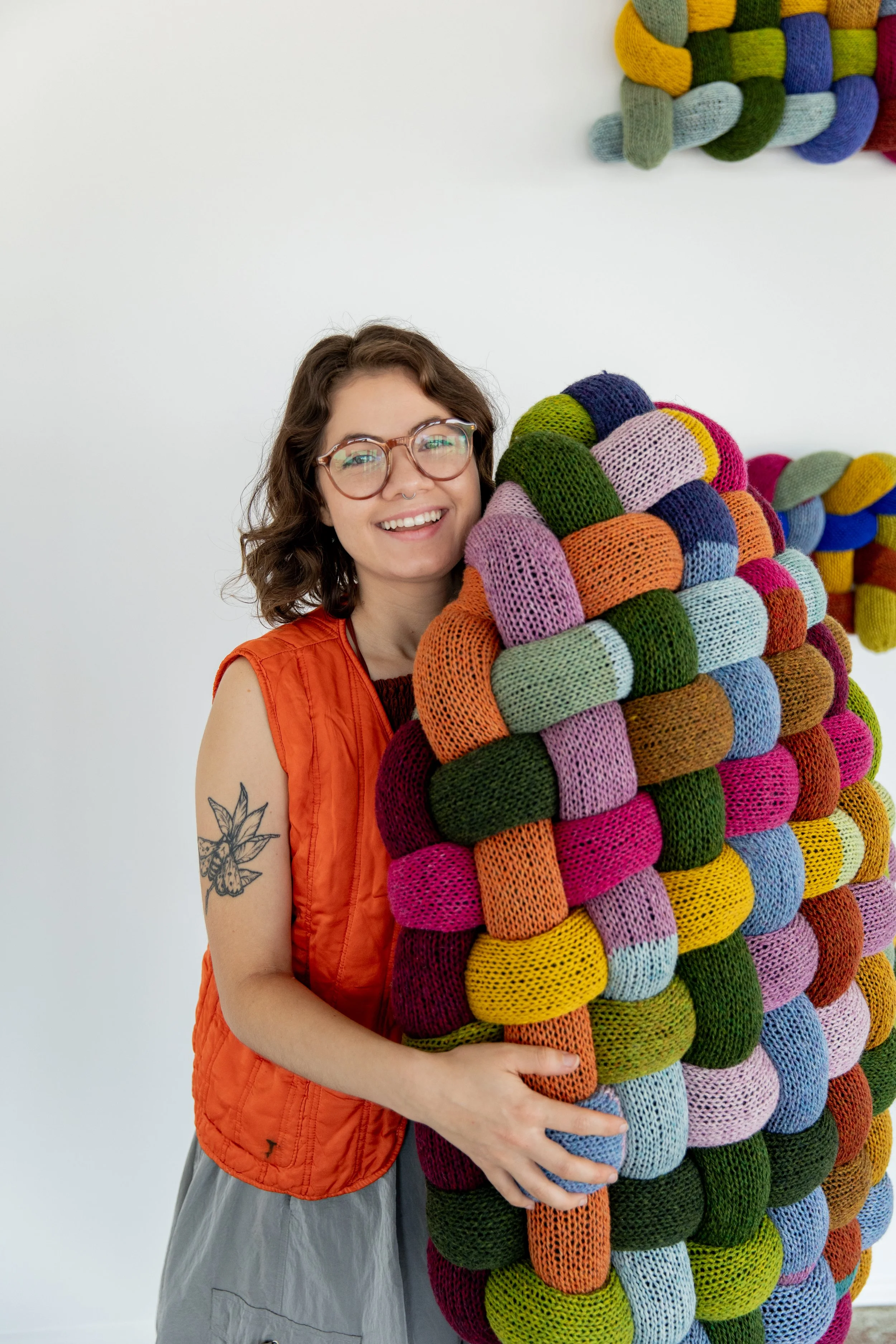 A woman with glasses, a tattoo on her arm, and a big smile, holding a colorful knitted sculpture made of many small knitted pieces in various colors including orange, pink, green, purple, and blue, against a plain white background.
