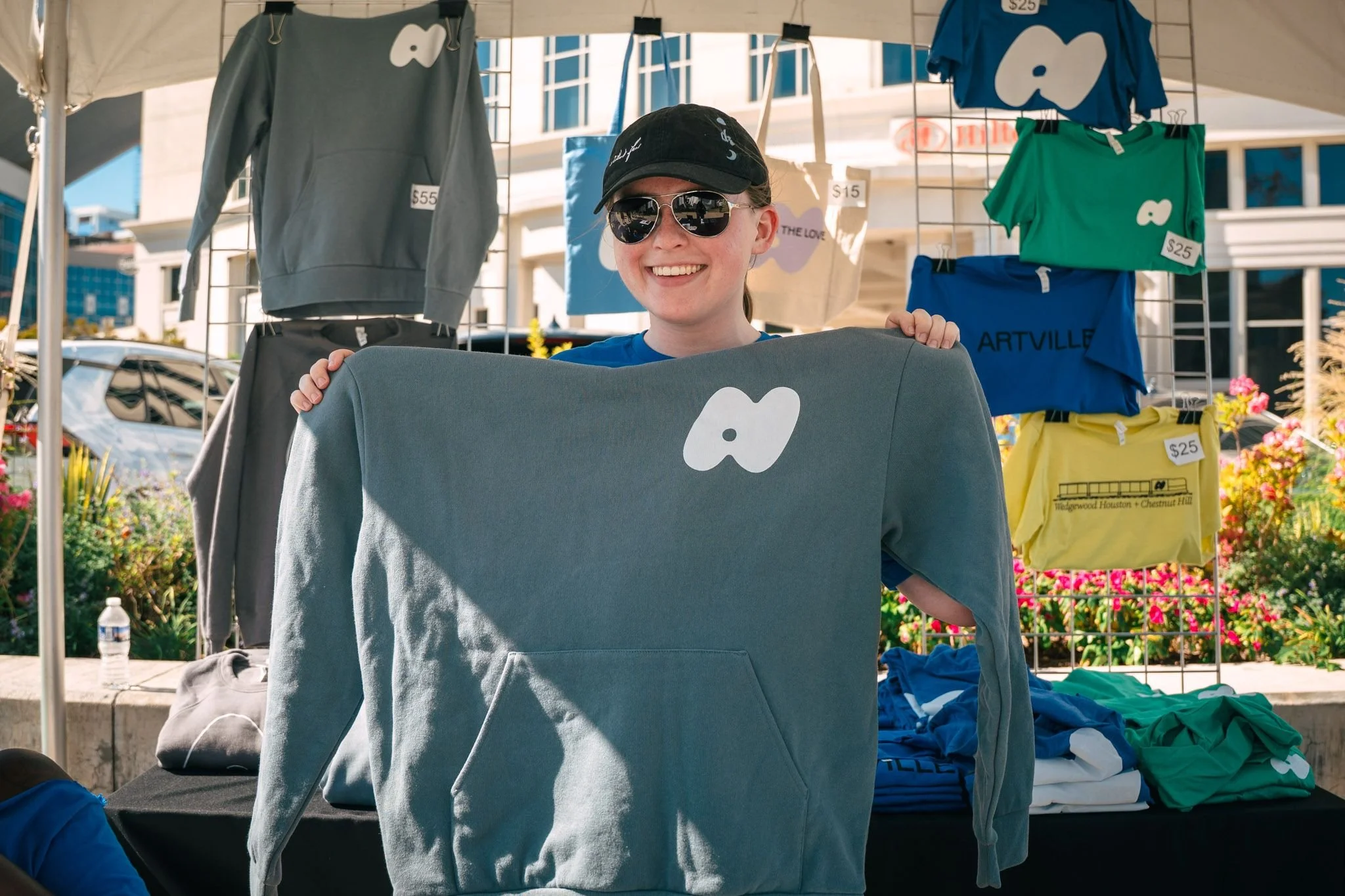 A smiling woman wearing sunglasses, a black cap, and a blue shirt, standing behind a gray hoodie with a white logo on it at an outdoor market stall with clothing on display.