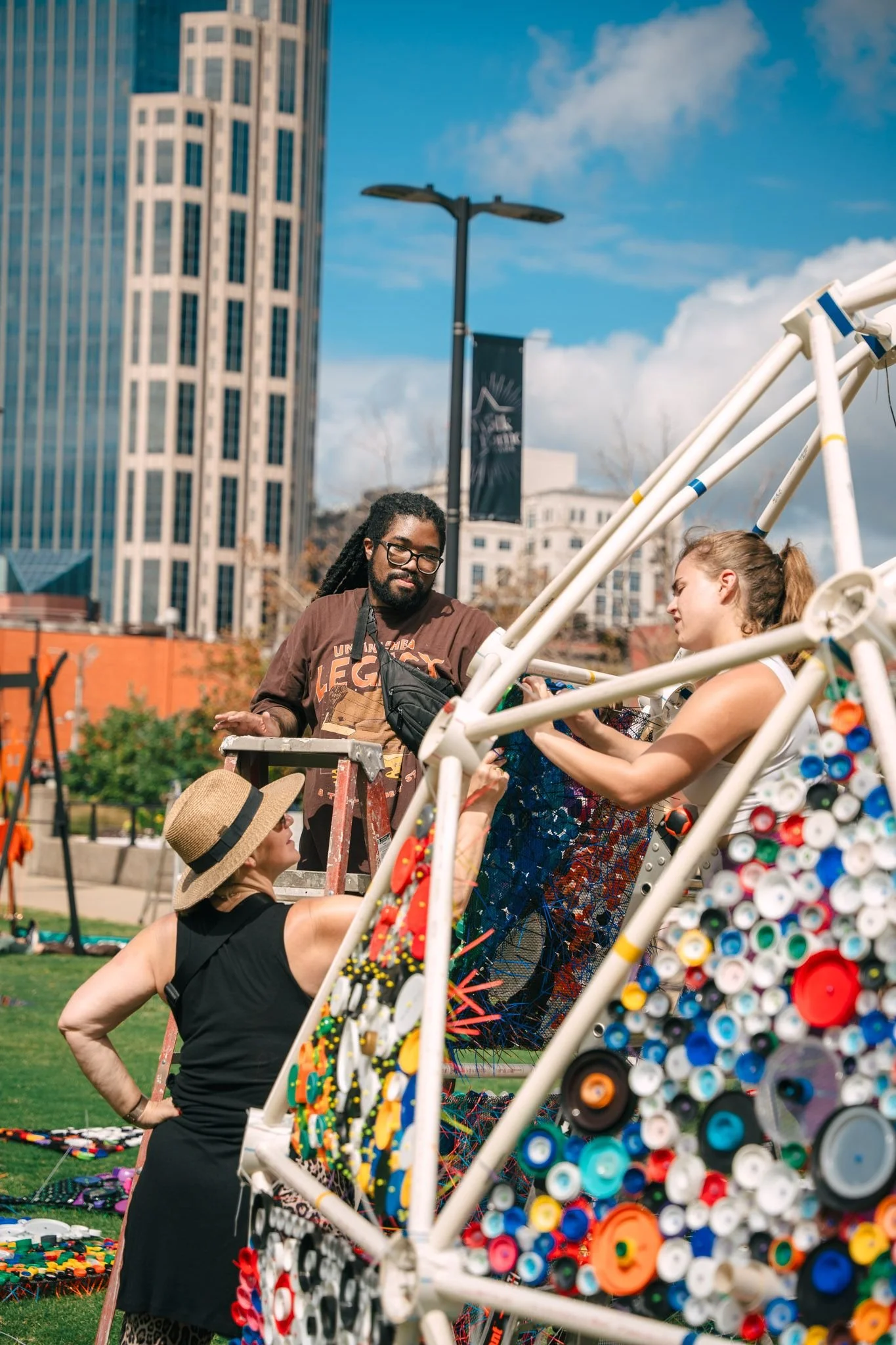 People at an outdoor art or craft fair with a large, colorful sculpture made of plastic bottle caps in the foreground, tall buildings and a blue sky in the background.