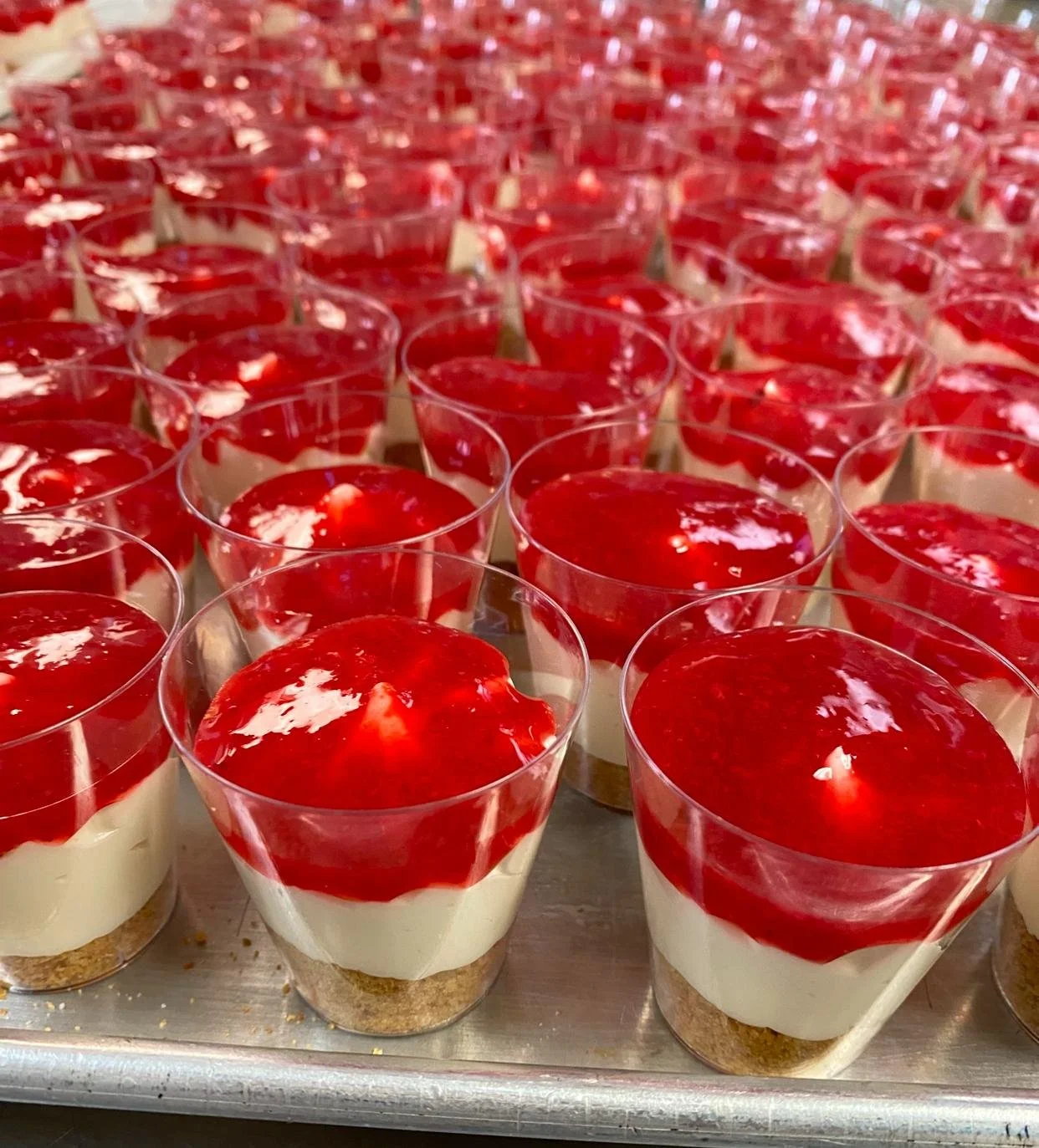 Multiple individual layered desserts with a crumb base, creamy middle layer, and red fruit topping, arranged in clear cups.  | Any Reason To Bake