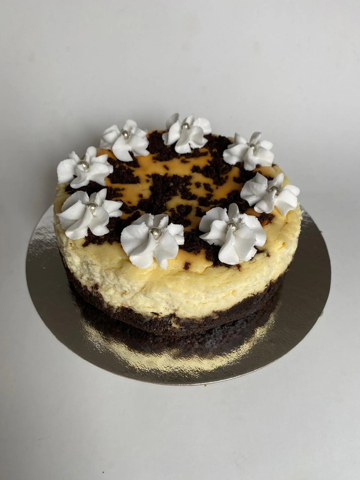 Cheesecake topped with white flowers, silver ball decorations, chocolate crumbles, and a caramel drizzle, placed on a silver cake board against a plain background. | Any Reason To Bake