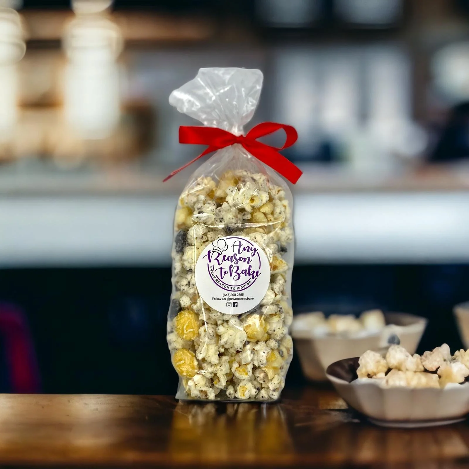 A clear plastic bag filled with popcorn, tied with a red ribbon. The bag has a white circular label that reads 'Any Reason to Bake' with contact information and social media icons. In the background, there are blurred shelves and bowls of popcorn.