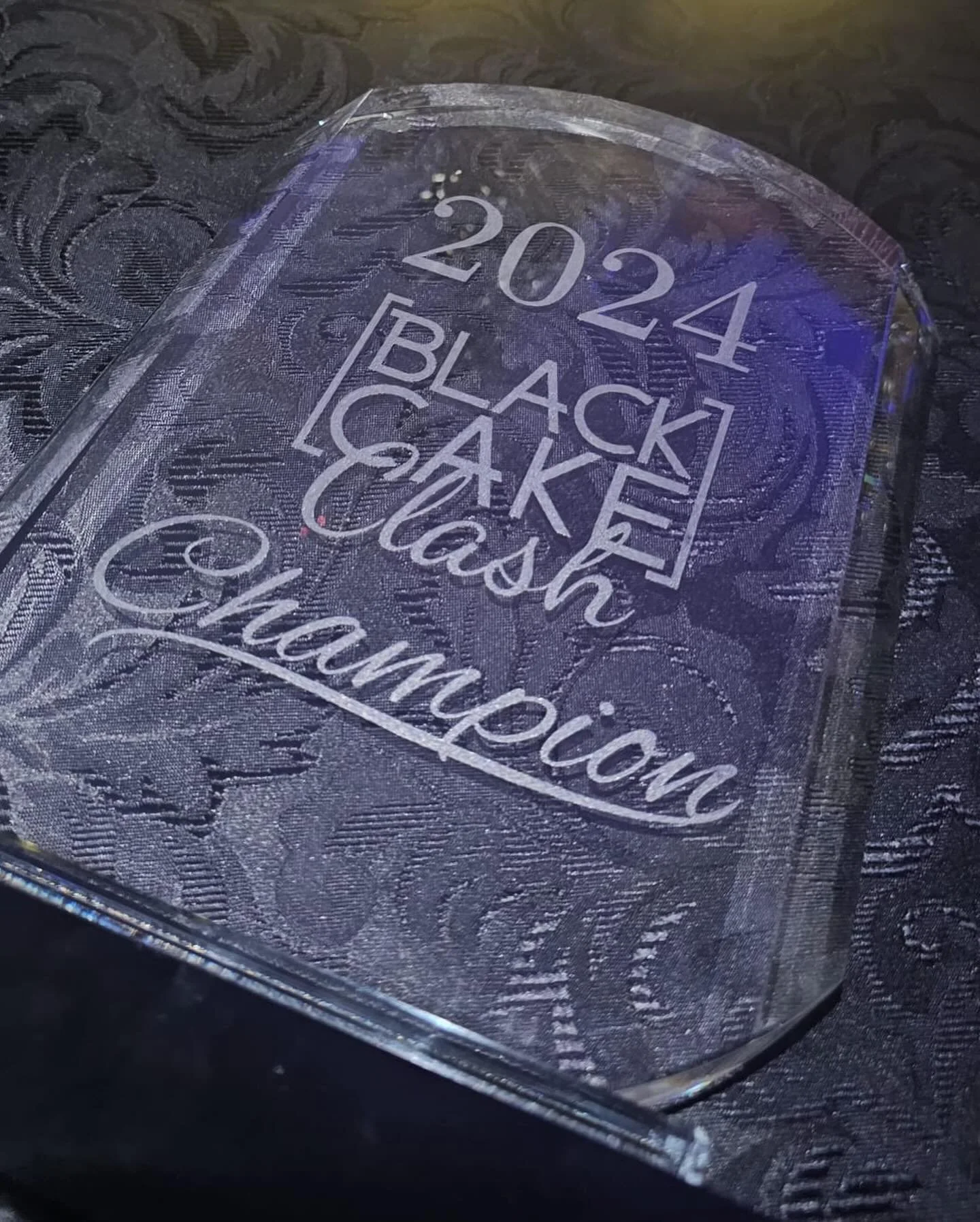 A clear glass awards with the inscription '2024 BLACK CAKE Championship' etched on it, placed on a black patterned surface. | Any Reason To Bake
