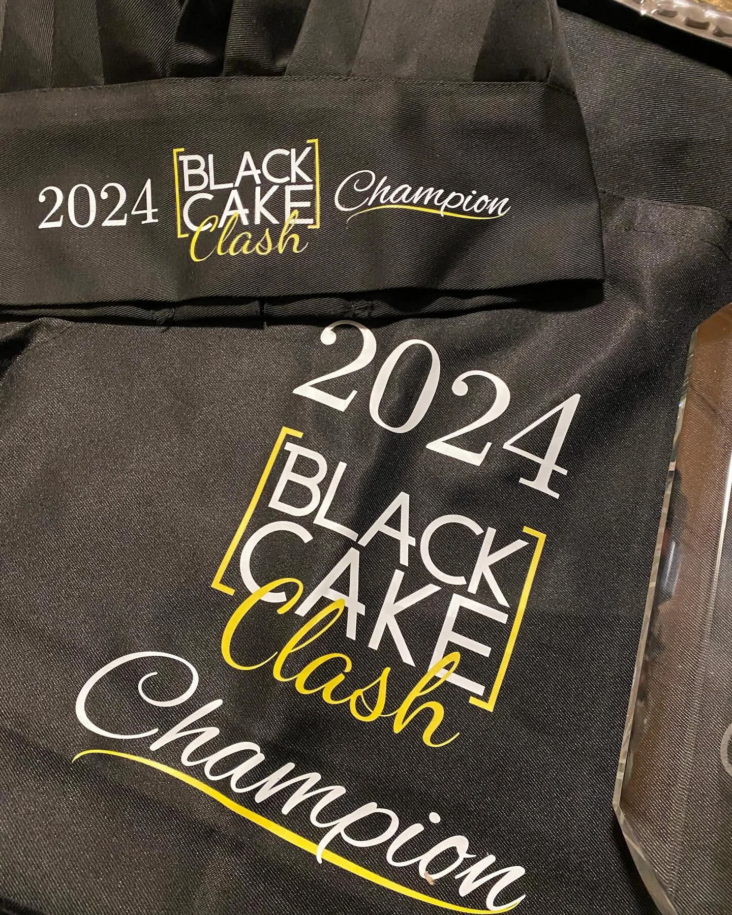 Black tote bag with text reading '2024 Black Cake Clash Champion' in white and yellow, placed among other similar bags. | Any Reason To Bake