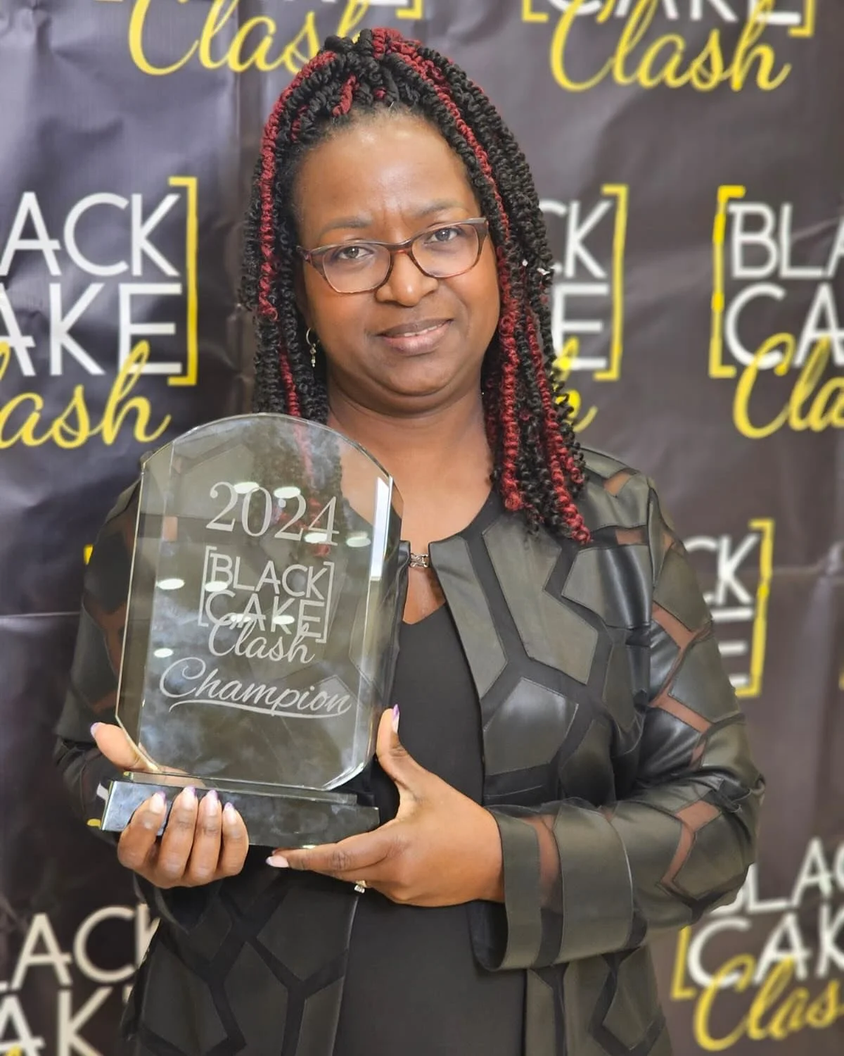 Woman holding a clear trophy with '2024 Black Cake Clash Champion' engraved on it, standing in front of a backdrop with 'Black Cake Clash' logos. | Any Reason To Bake
