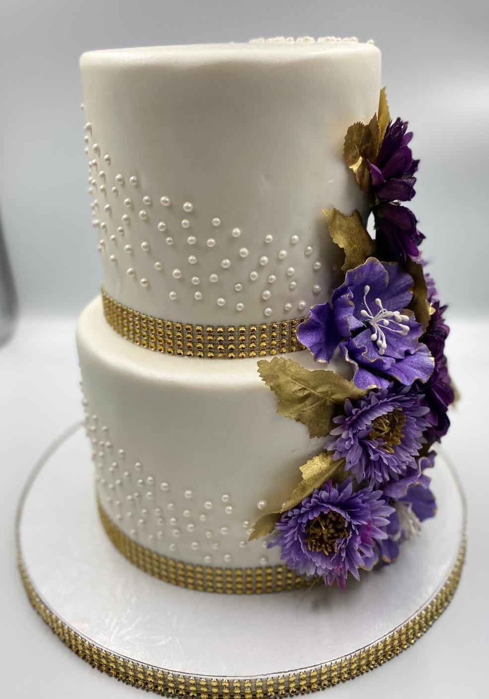 Two-tier white wedding cake decorated with purple flowers, gold leaves, pearl-like beads, and gold ribbon accents.