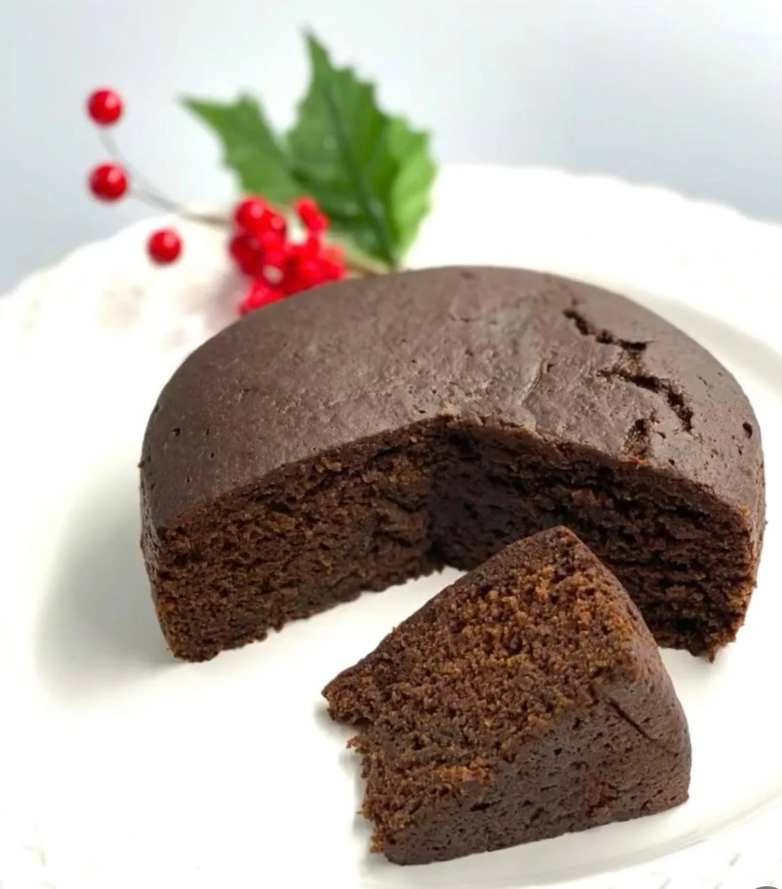 Chocolate cake with a slice cut out, garnished with holly leaves and red berries, served on a white plate.